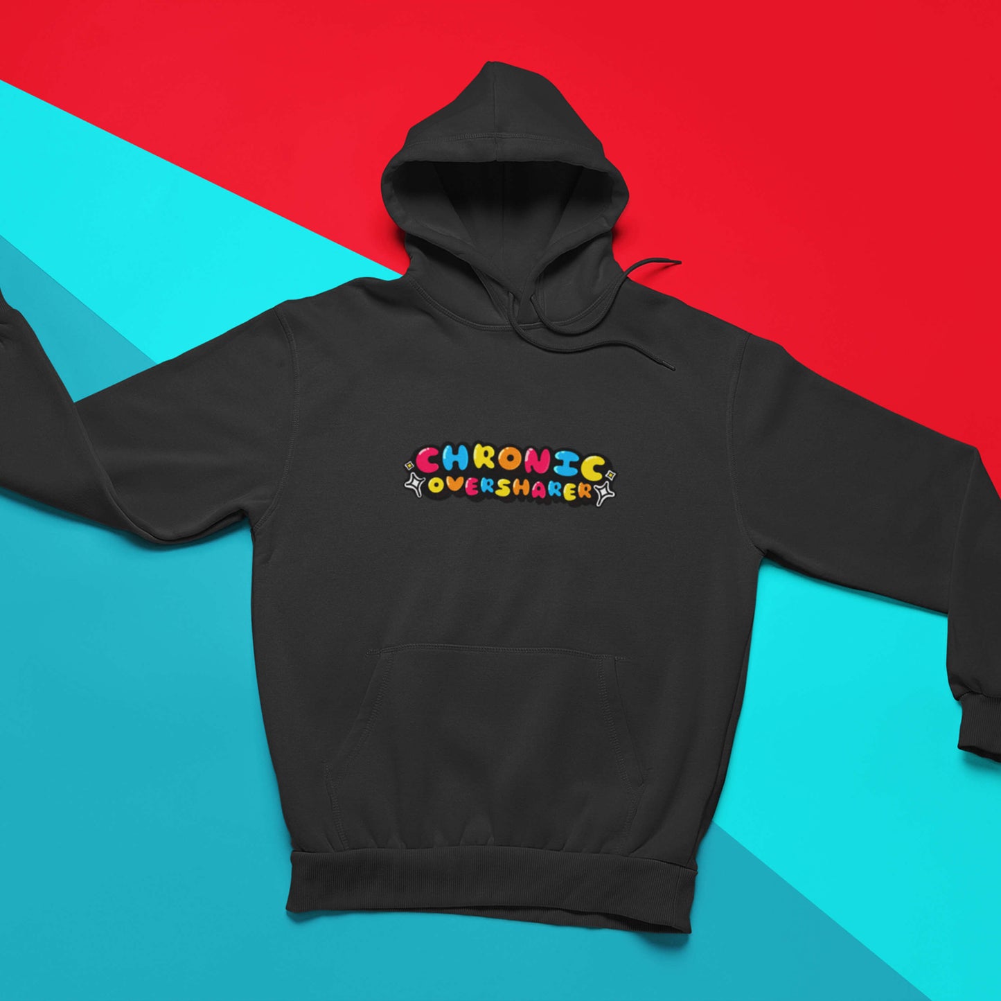 The Chronic Over Sharer Black Hoodie laying on a red and blue background. The black hoodie features a drawstring hood, a large front pocket and the text 'chronic oversharer' in rainbow bubble font with a black shadow effect and white sparkles. The design was created to raise awareness for neurodivergent disorders such as ADHD.