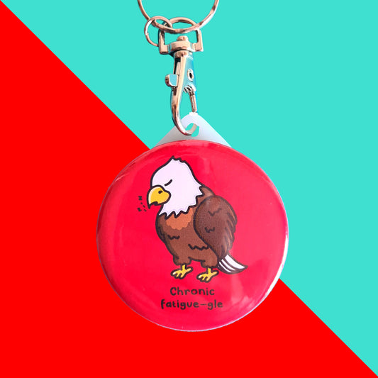 The Chronic Fatigue-gle Keyring - Chronic Fatigue on a blue and red background. The silver metal clip with plastic circular keychain is a red base with a sleeping tired brown and white eagle with a yellow beak and feet, underneath in black text reads 'chronic fatigue-gle'. The design was created to raise awareness for chronic fatigue / ME / CFS.