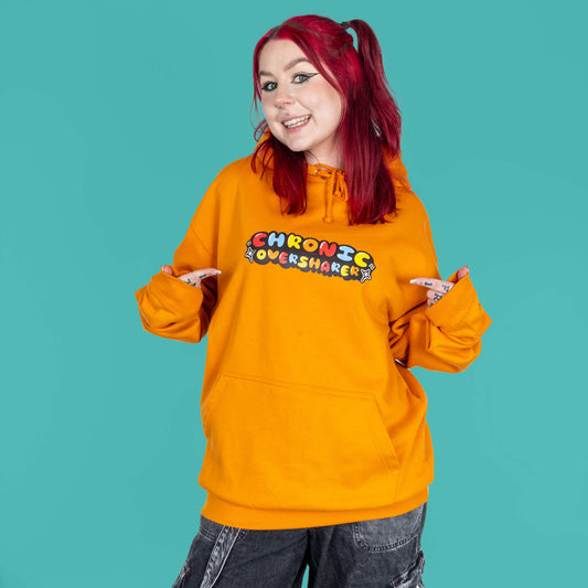 The Chronic Over Sharer Pumpkin Orange Hoodie modelled on Flo, a red haired alternative model, in front of a blue background. She is facing forward smiling pointing towards the middle of the hoodie, photo is cropped from the thighs up. The pumpkin orange hoodie features a drawstring hood, a large front pocket and the text 'chronic oversharer' in rainbow bubble font with a black shadow effect and white sparkles. The design was created to raise awareness for neurodivergent disorders such as ADHD.