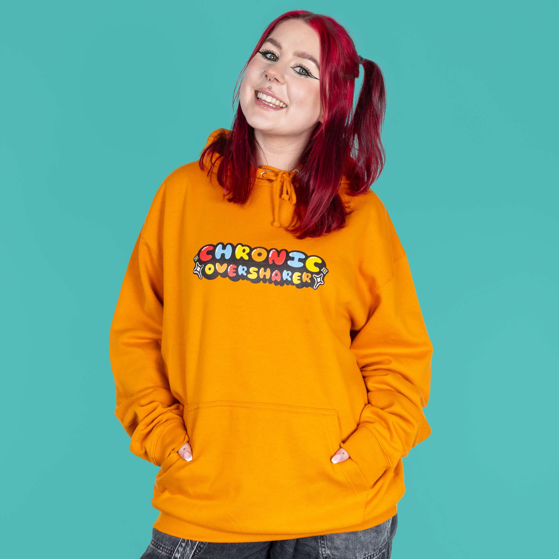 The Chronic Over Sharer Pumpkin Orange Hoodie modelled on Flo, a red haired alternative model, in front of a blue background. She is facing forward smiling with both hands in the front pocket, photo is cropped from the thighs up. The pumpkin orange hoodie features a drawstring hood, a large front pocket and the text 'chronic oversharer' in rainbow bubble font with a black shadow effect and white sparkles. The design was created to raise awareness for neurodivergent disorders such as ADHD.
