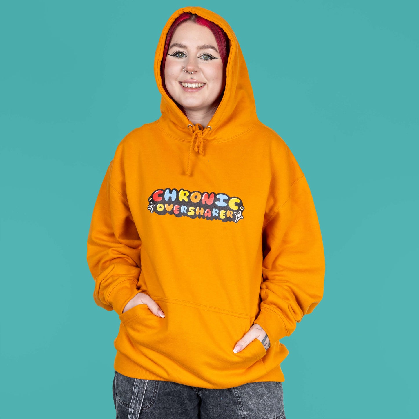The Chronic Over Sharer Pumpkin Orange Hoodie modelled on Flo, a red haired alternative model, in front of a blue background. She is facing forward smiling with both hands in the front pocket and the hood up, photo is cropped from the thighs up. The pumpkin orange hoodie features a drawstring hood, a large front pocket and the text 'chronic oversharer' in rainbow bubble font with a black shadow effect and white sparkles. The design was created to raise awareness for neurodivergent disorders such as ADHD.