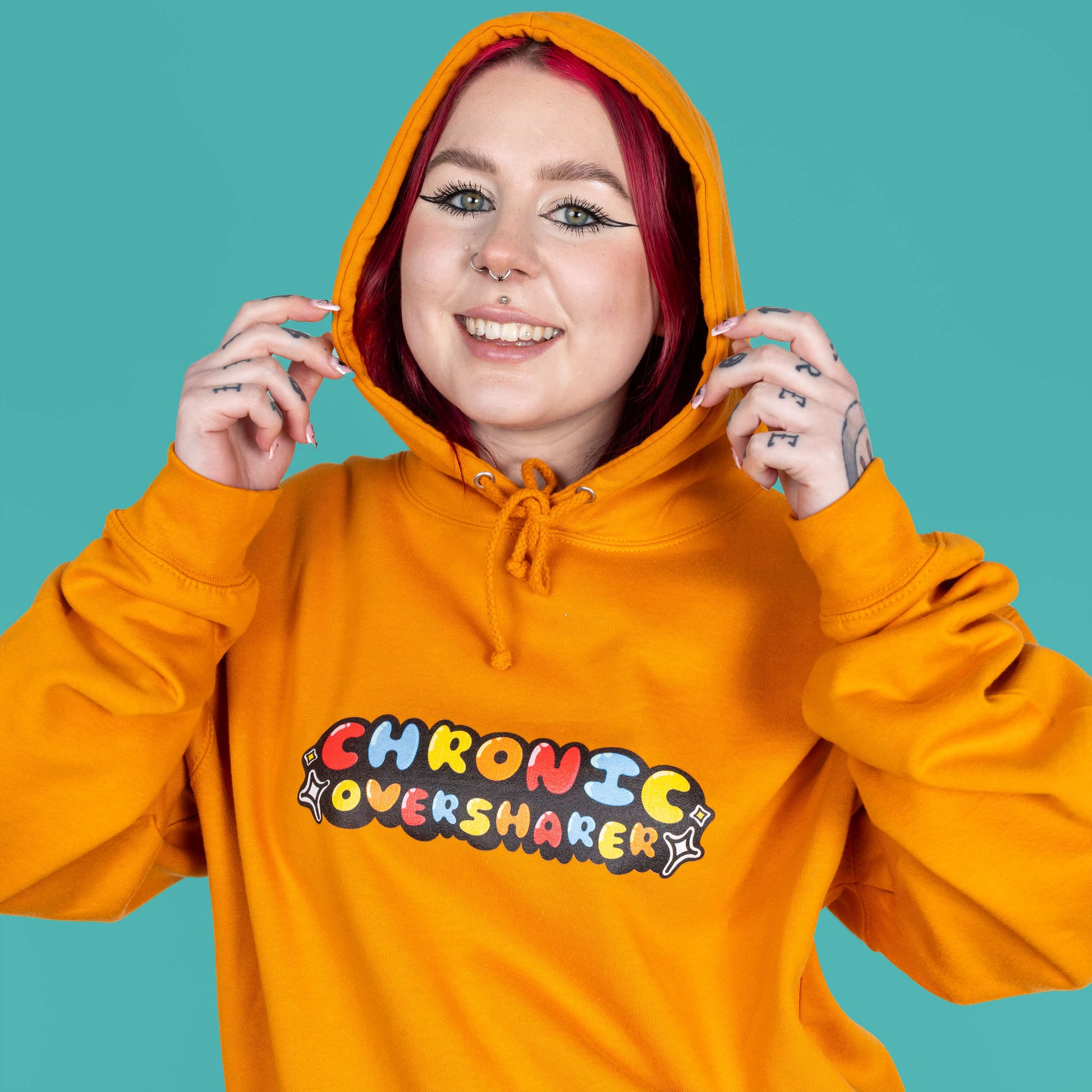 The Chronic Over Sharer Pumpkin Orange Hoodie modelled on Flo, a red haired alternative model, in front of a blue background. She is facing forward smiling with both hands resting on the hood up, photo is cropped from the waist up. The pumpkin orange hoodie features a drawstring hood, a large front pocket and the text 'chronic oversharer' in rainbow bubble font with a black shadow effect and white sparkles. The design was created to raise awareness for neurodivergent disorders such as ADHD.