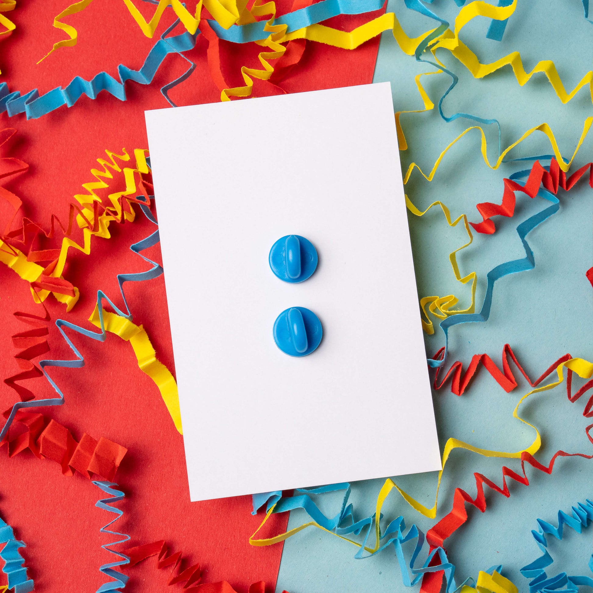 Back view of Chronic Pain guin penguin enamel pin by Innabox showing two blue rubber pin backs attached to a white card. The enamel pin fastenings are clearly visible. Styled on a colourful red and blue background with yellow, blue and red paper streamers.
