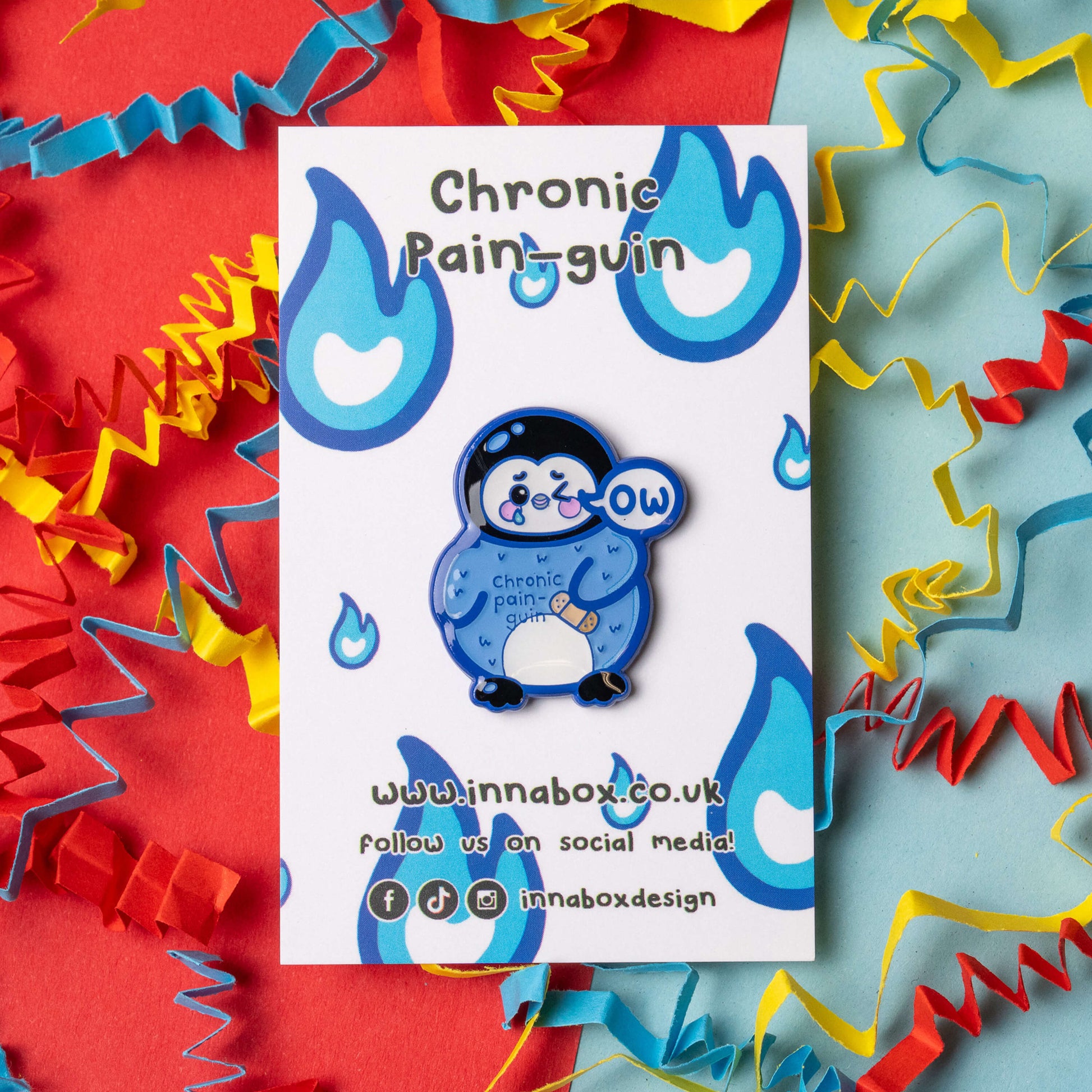Chronic Pain guin penguin enamel pin by Innabox shown on white branded backing card. Blue penguin character with an ow speech bubble, blushing cheeks and a small plaster illustration. The backing card features blue flame style graphics and Innabox branding. Bright colourful streamer background in red, blue and yellow.
