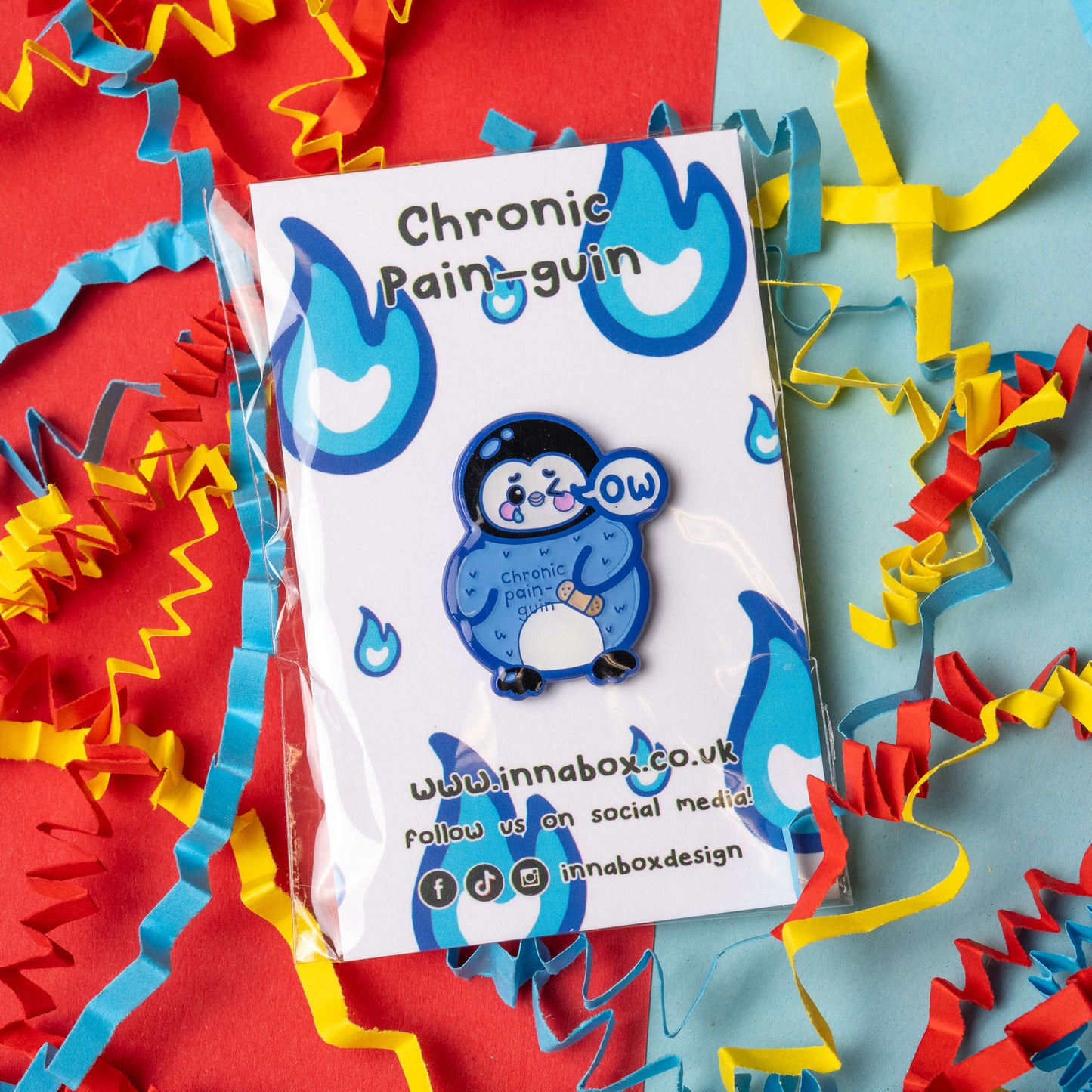 Chronic Pain guin penguin enamel pin by Innabox displayed on branded backing card. Cute blue penguin character with teary eyes and a speech bubble saying ow. The penguin has a plaster on its belly and the words chronic pain guin on its chest. Colourful background with red, blue and yellow paper streamers.