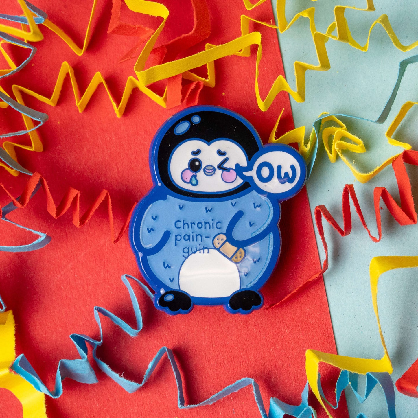 Close up of the Chronic Pain guin penguin enamel pin by Innabox. Blue penguin character with a sad but cute expression, ow speech bubble and a plaster on its tummy. The words chronic pain guin are visible on the penguin. Photographed on a bright red and blue background with colourful paper streamers.