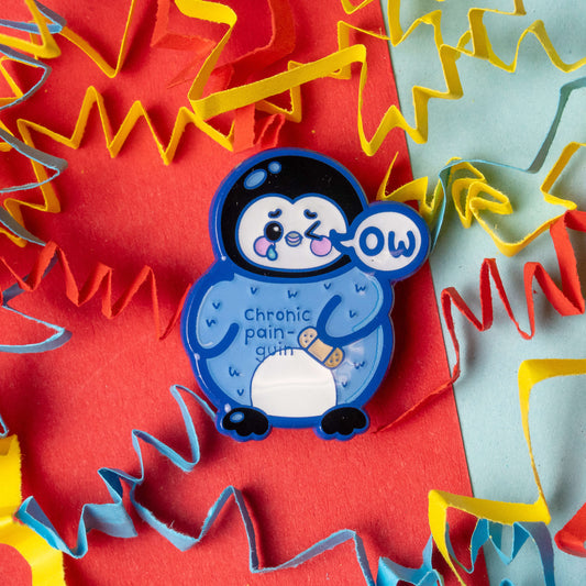 Close up of the Chronic Pain guin penguin enamel pin by Innabox. Blue penguin character with a sad but cute expression, ow speech bubble and a plaster on its tummy. The words chronic pain guin are visible on the penguin. Photographed on a bright red and blue background with colourful paper streamers.