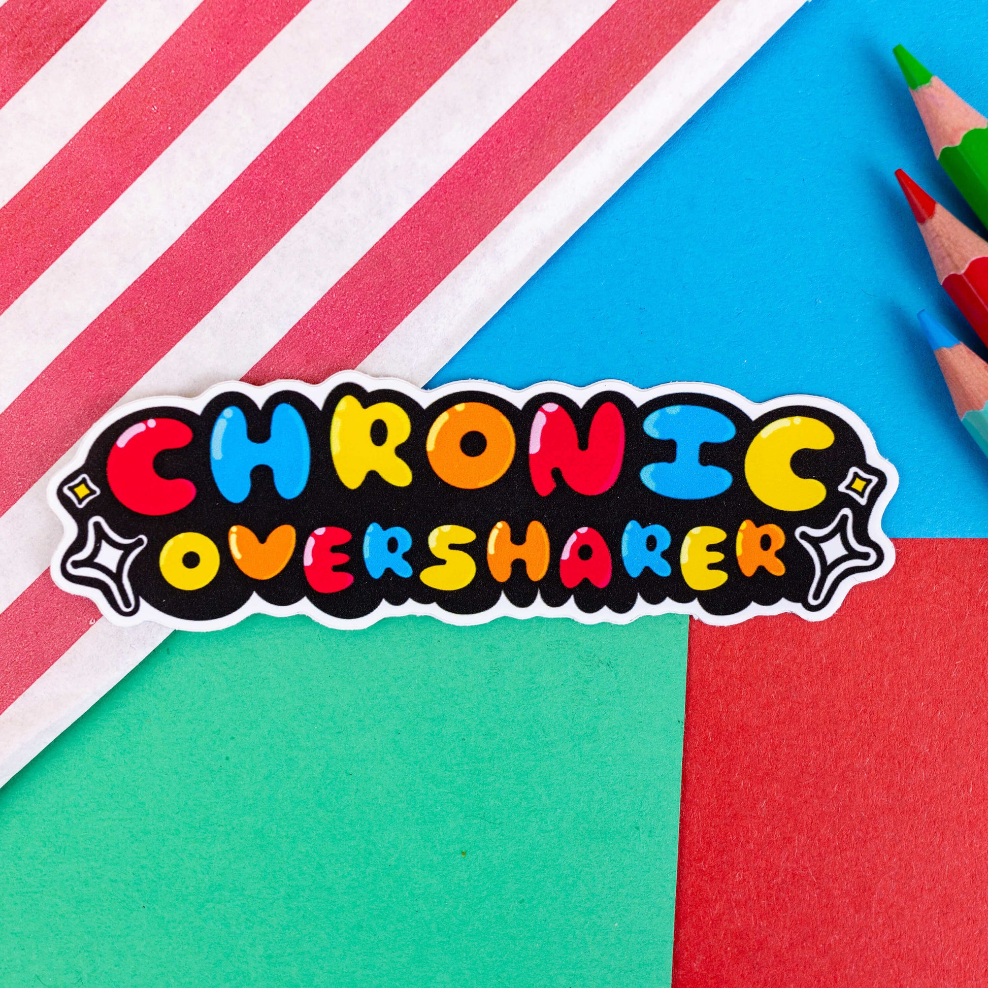 The Chronic Oversharer Sticker on a blue, green and red background with colouring pencils and a red stripe candy bag. The sticker is rainbow bubble font reading 'chronic oversharer' with a black drop shadow and white sparkles surrounding. The sticker was designed to raise awareness of neurodivergent disorders such as ADHD.