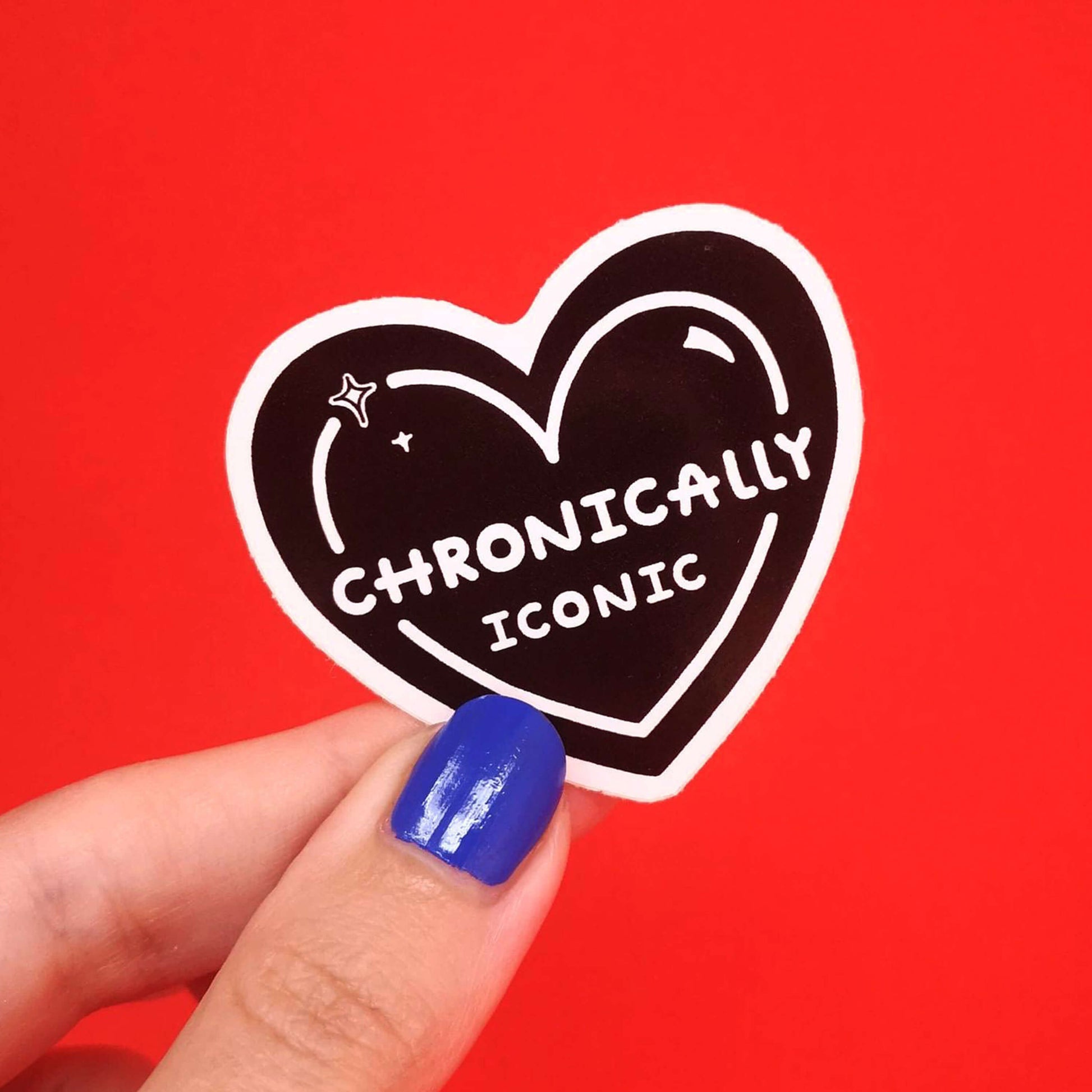 The Chronically Iconic Sticker being held over a red background by a hand with blue nail varnish. The black heart shaped sticker has a white outline with sparkles and centre text reading 'chronically iconic'. The design is raising awareness for chronic illness and invisible illness.