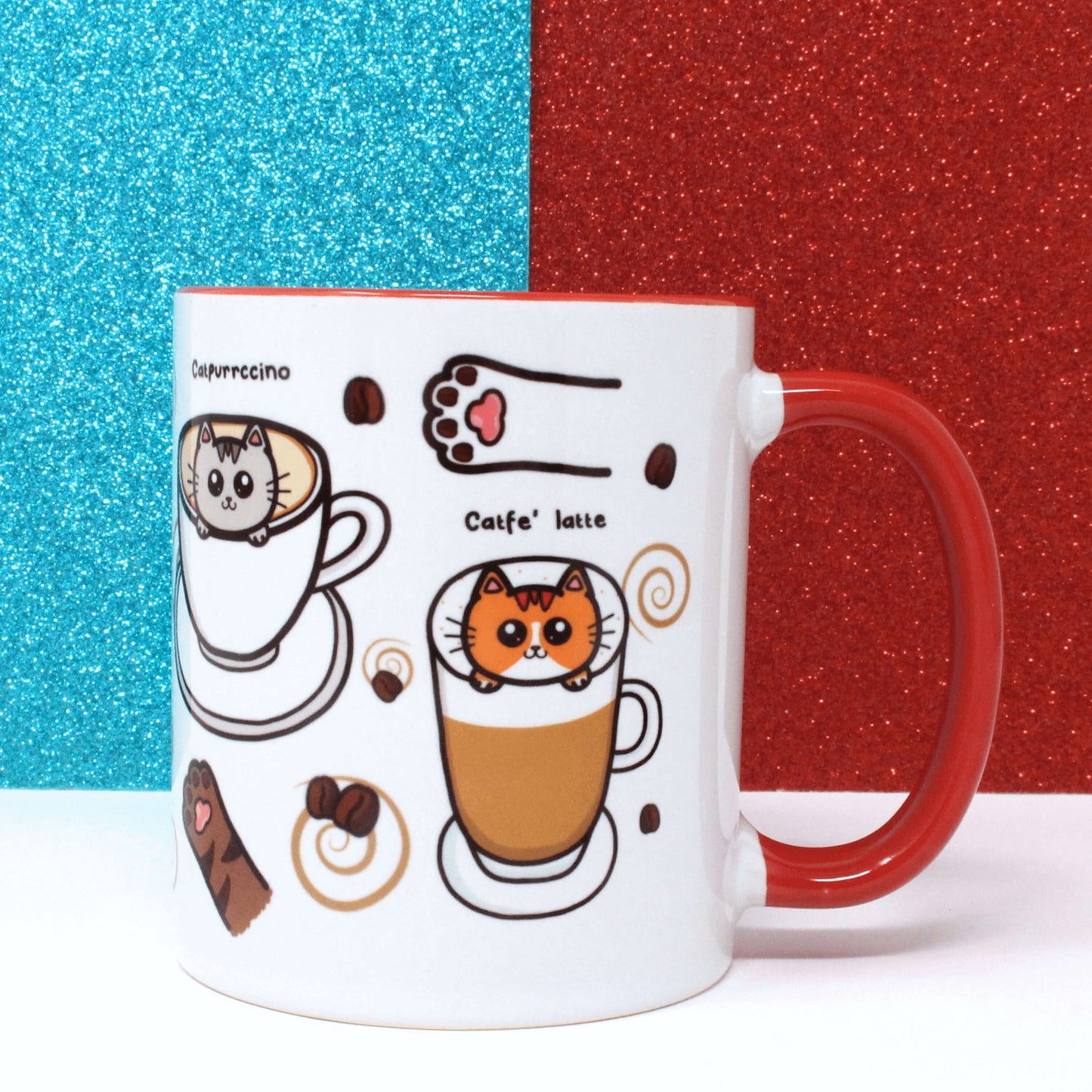 The Coffee Mog Cat Mug on a white table with a red and blue glitter background. The cat themed white coffee mug with red inner and handle is facing left to show illustrations of cat paw toe coffee beans, catpurrccino (a grey cat in a cappuccino), catfe' latte (an orange cat in a latte) with coffee beans and brown swirls.