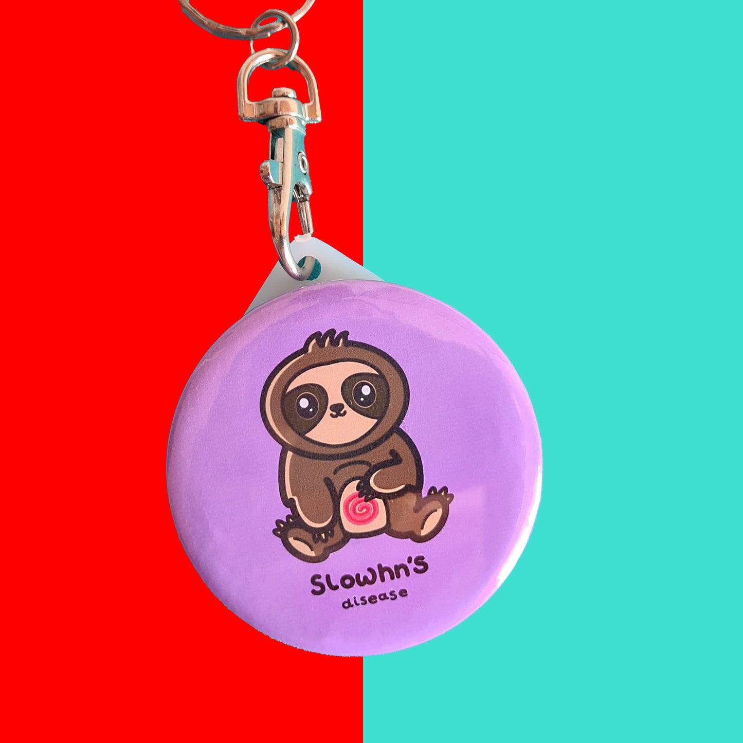 The Slowhn's Disease Sloth Keyring - Crohn's Disease on a red and blue background. The silver lobster clip pink plastic circular keyring of a smiling sat down brown sloth clutching its swirly red tummy and black text underneath reading 'slowhn's disease'. The hand made design is raising awareness for crohn's disease.