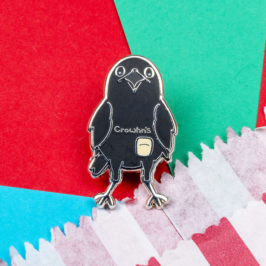 The Crowhn's Disease Enamel Pin - Crohn's Disease on a red, blue and teal background next to a red and white striped paper bag. The black crow shape pin has a white stoma bag fitted and the text reading 'chrowhn's' across its chest. The design is raising awareness for crohn's disease.