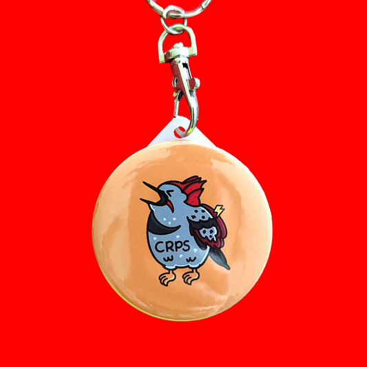 CRPS Keyring - Complex Regional Pain Syndrome on a red background. The silver clip yellow plastic circular keychain features a screaming upset woodpecker bird with a lightning bolt on its wing and 'CRPS' written across its belly in black. The design is raising awareness for complex regional pain syndrome.