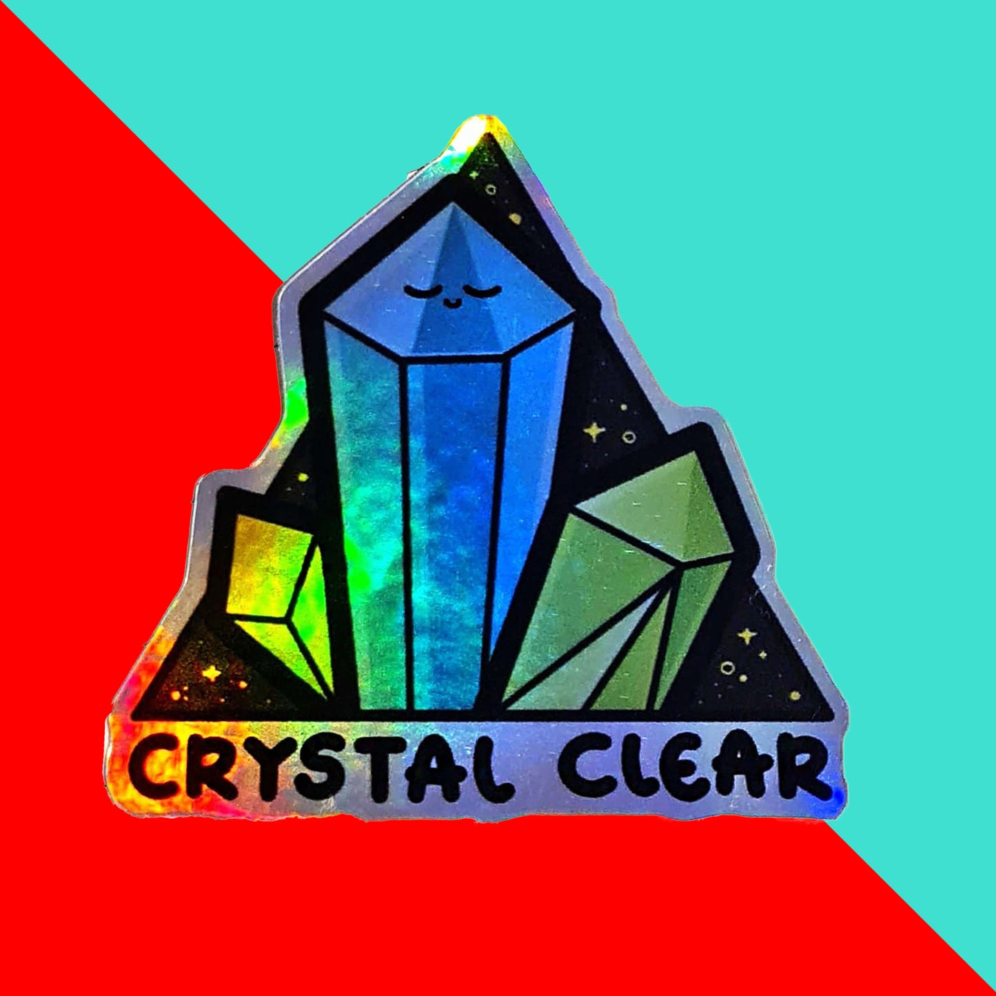 The Crystal Clear Holographic Sticker on a red and blue background. The triangle shaped sticker features three green crystal towers on a black sparkly background with bottom text reading crystal clear, the centre crystal is smiling. Inspired by witchy spiritual healing with crystals.