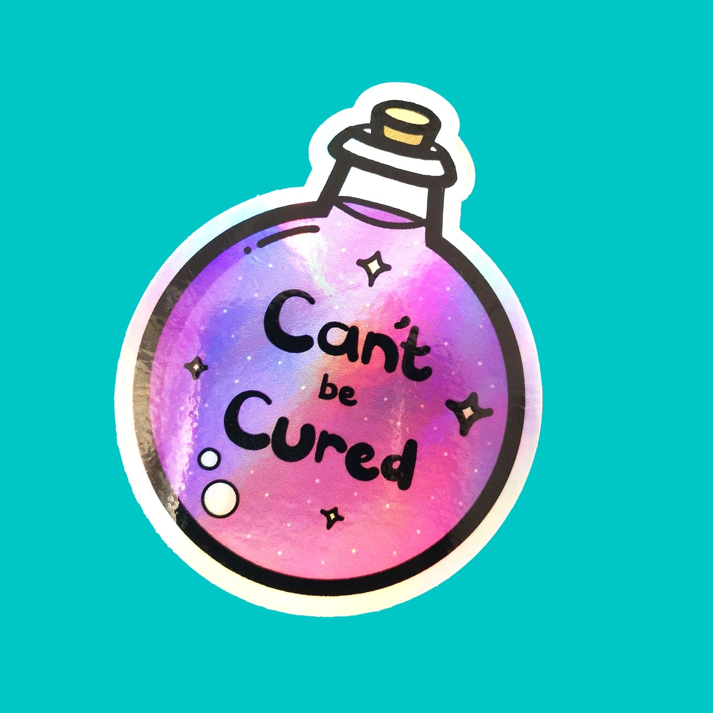 The Can't Be Cured Holographic Sticker on a blue background. The sticker is a circular potion bottle with a cork stop top and purple middle. In the middle is white and yellow sparkles, two pastel pink bubbles and black text that reads 'can't be cured'. The design was inspired by chronic illnesses.