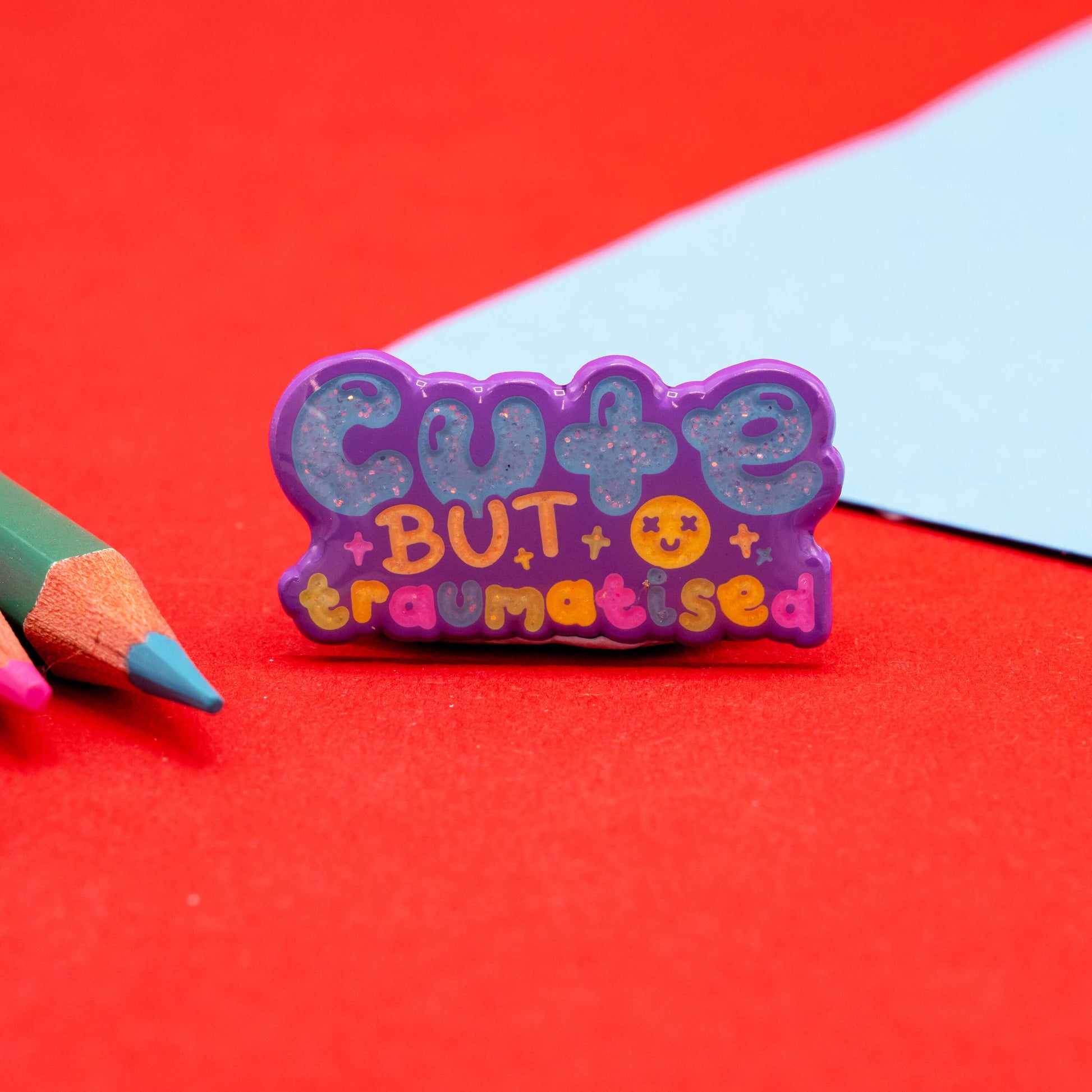 Glittery “Cute But Traumatised” enamel pin by Innabox in pastel colours with a purple border, styled on red paper with coloured pencils and a pale blue background. Disability awareness accessory for jackets, bags, and more.