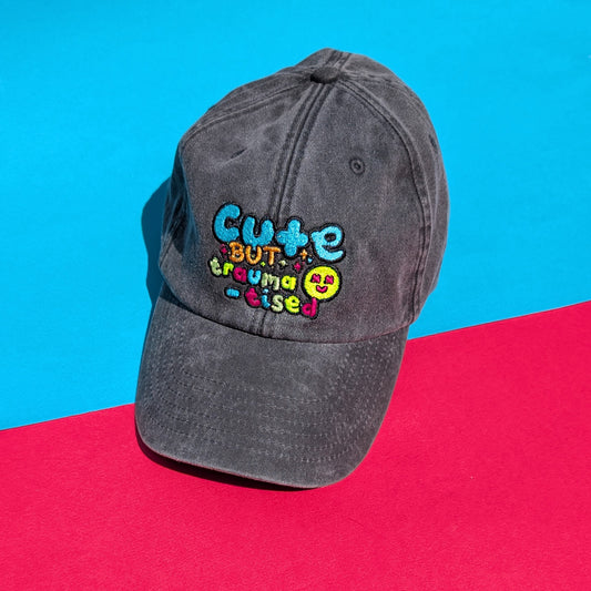 Flat lay of the Innabox Hot and Neuro-Spicy cap on a bright red and blue background, showcasing the embroidered “Cute but traumatised” text on vintage black-grey brushed cotton.