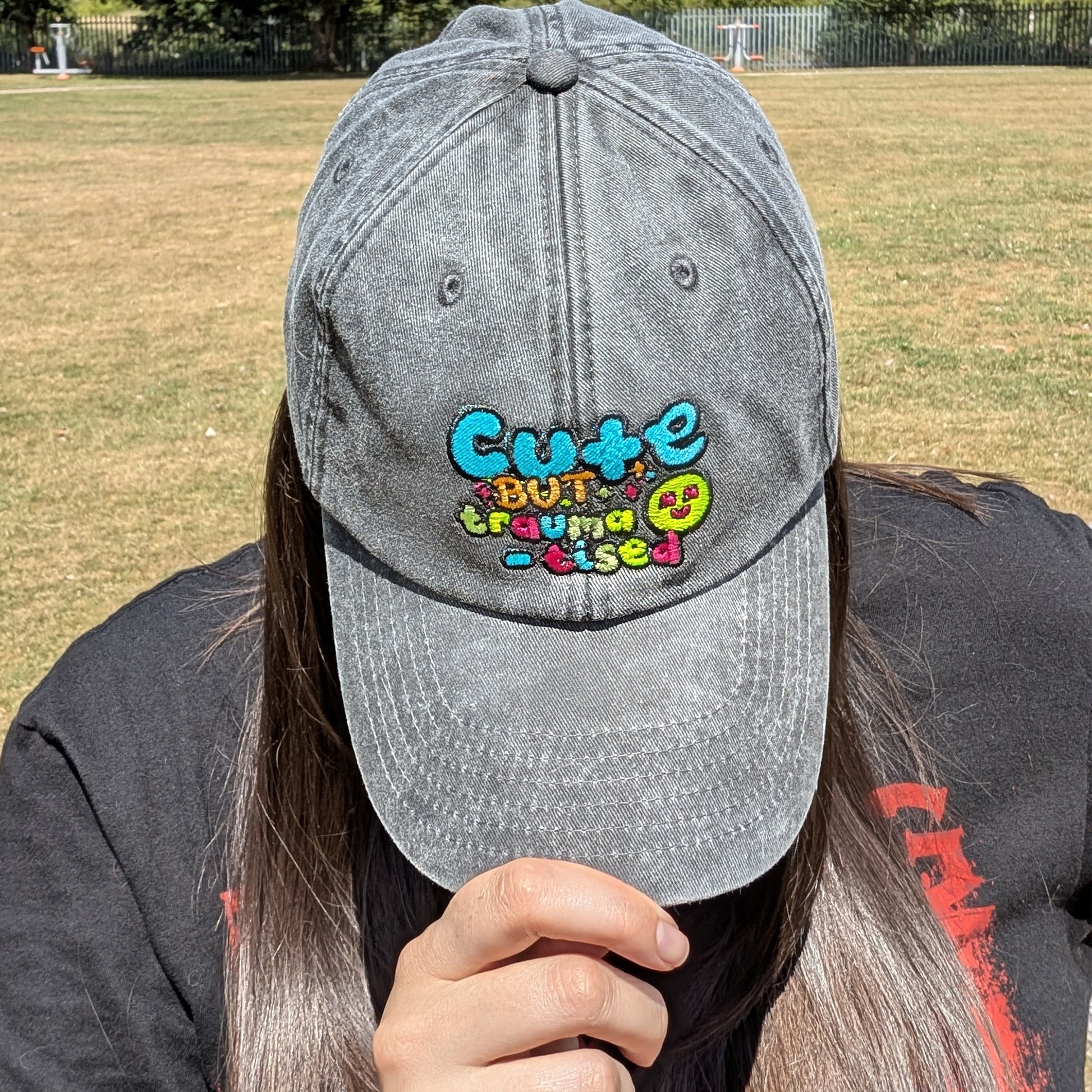Model wearing the Hot and Neuro-Spicy cap by Innabox, showing the embroidered slogan "Cute but traumatised" from a top-down angle while seated in a sunny park.