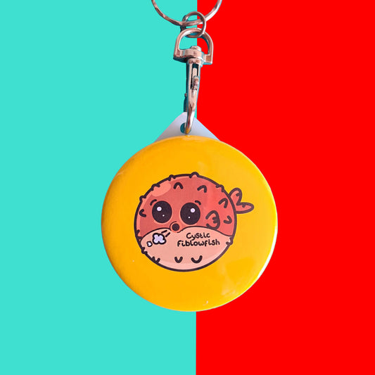 The Cystic Fiblowfish Keyring - Cystic Fibrosis on a red and blue background. The silver clip plastic yellow circle keychain with a puffer fish blowfish wheezing with 'cystic fiblowfish' written in black on its belly. The design is raising awareness for cystic fibrosis.