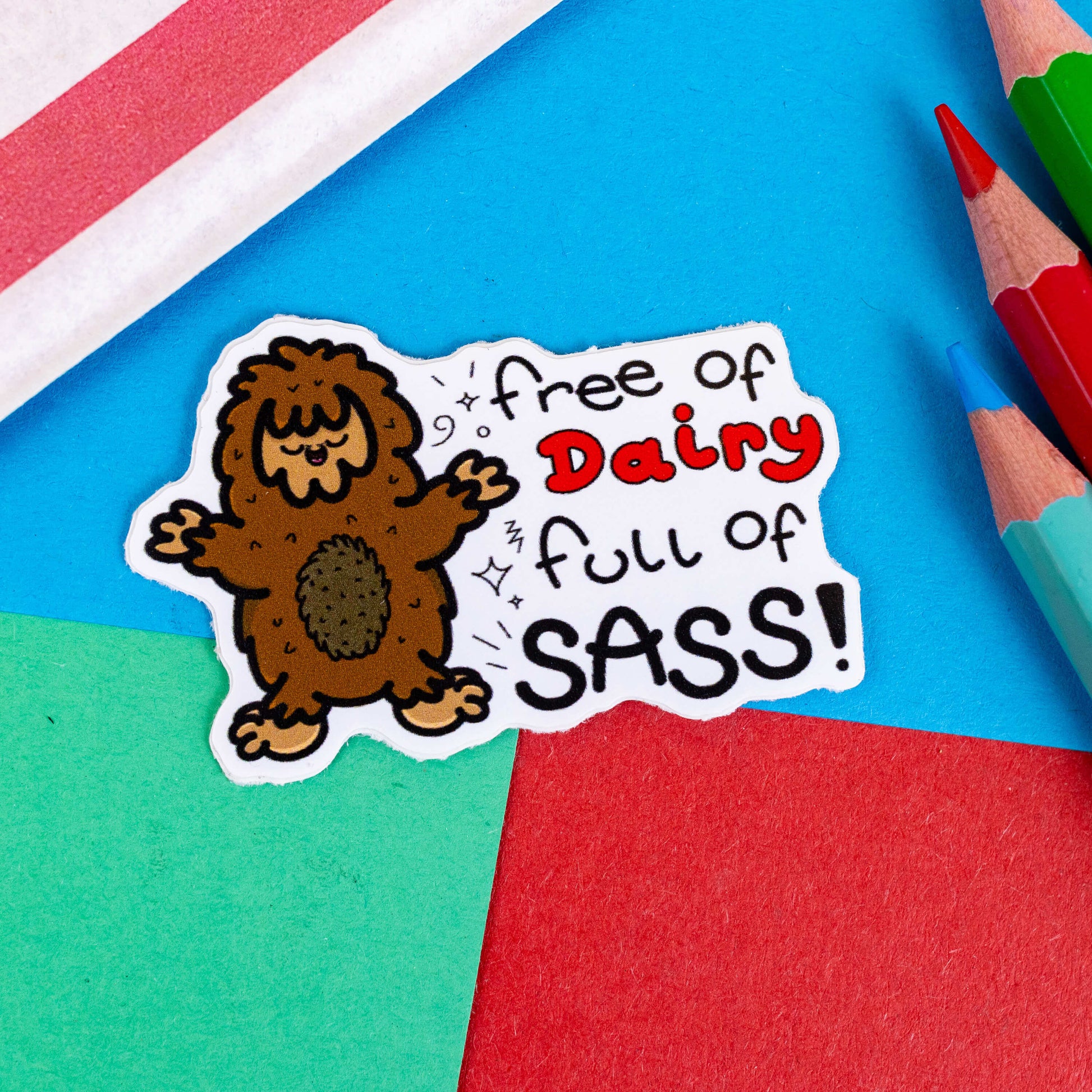 The Free of Dairy, Full of Sass! Sticker - Lactose Intolerant on a red, blue and green background with colouring pencils and a red stripe candy bag. The sticker features a smiling cheering brown sasquatch with black sparkles and text reading 'free of dairy full of sass!'. The design was created to raise awareness for lactose intolerance.