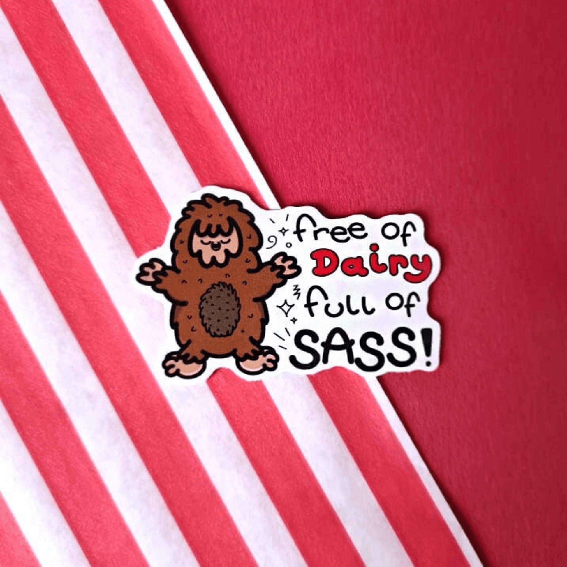 The Free of Dairy, Full of Sass! Sticker - Lactose Intolerant on a red background with a red stripe candy bag. The sticker features a smiling cheering brown sasquatch with black sparkles and text reading 'free of dairy full of sass!'. The design was created to raise awareness for lactose intolerance.