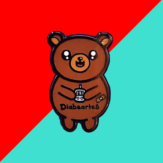 The Diabeartes Enamel Pin - Diabetes on a red and blue background. The brown bear shaped pin is smiling holding a blood glucose reader with a plaster on its arm, across its tummy reads 'diabeartes' in black. The design is raising awareness for those with type 1 diabetes and type 2 diabetes.