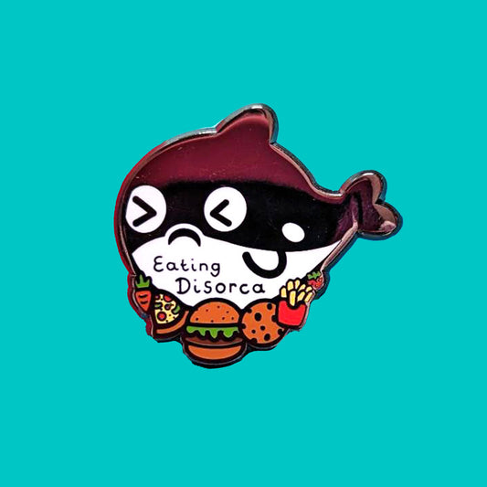 The Eating Disorca Orca Whale Enamel Pin - Eating Disorder on a blue background. The black orca whale shaped enamel pin has a stressed expression whilst surrounded by burgers, pizzas, cookies, fries and fruit with 'eating discorca' across its middle. The design is raising awareness for eating disorders.
