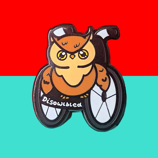 The Disowlbled Enamel Pin - Disabled on a red and blue background. A brown tawny owl with yellow eyes sat on a silver and black wheelchair that has white text reading 'disowlbled'. The design is raising awareness for disabilities and invisible illnesses.