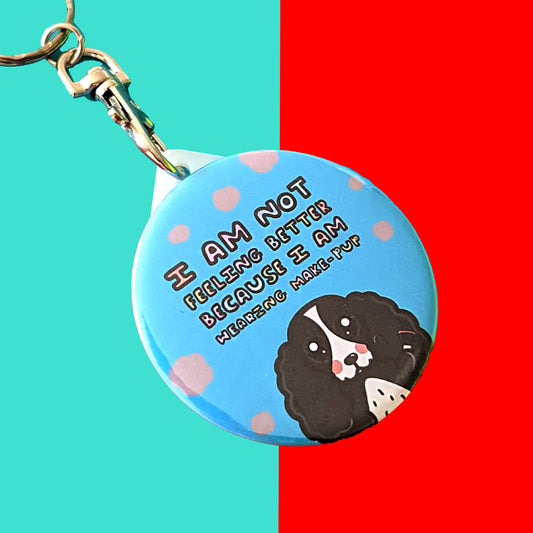 I Am Not Feeling Better - Dog Keyring on a red and blue background. The blue circular keyring has white text with black outline reading 'I am not better because I am wearing make-up'. There is an illustration of a black and white dog underneath the text with big sparkly eyes and rosy cheeks. The hand drawn design is made to raise awareness for invisible and chronic illnesses.