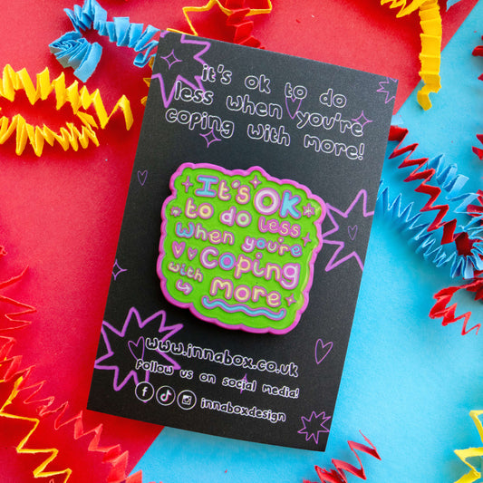 Close up of Innabox enamel pin reading “It’s OK To Do Less When You’re Coping With More” on black backing card with purple doodles. Bright green, pink and pastel lettering. Styled on colourful red and blue background with decorative streamers. Positive mental health and disability awareness accessory.
