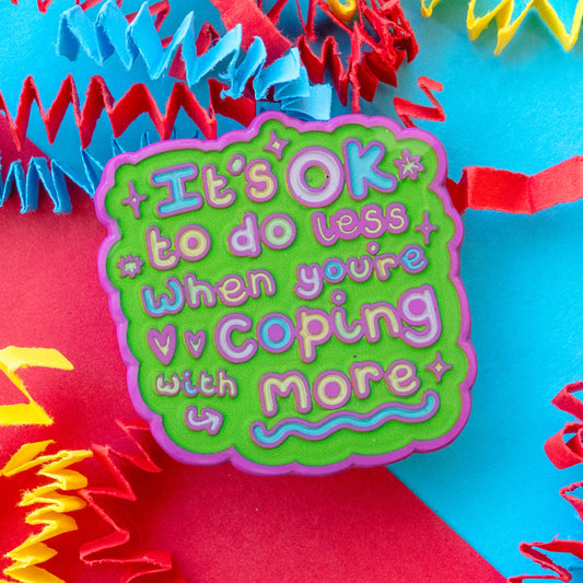 Close up of “It’s OK To Do Less When You’re Coping With More” enamel pin by Innabox. Bright green design with pink border and pastel lettering. Styled on bold red and blue background with paper streamers. Uplifting accessory supporting chronic illness and hidden disabilities.