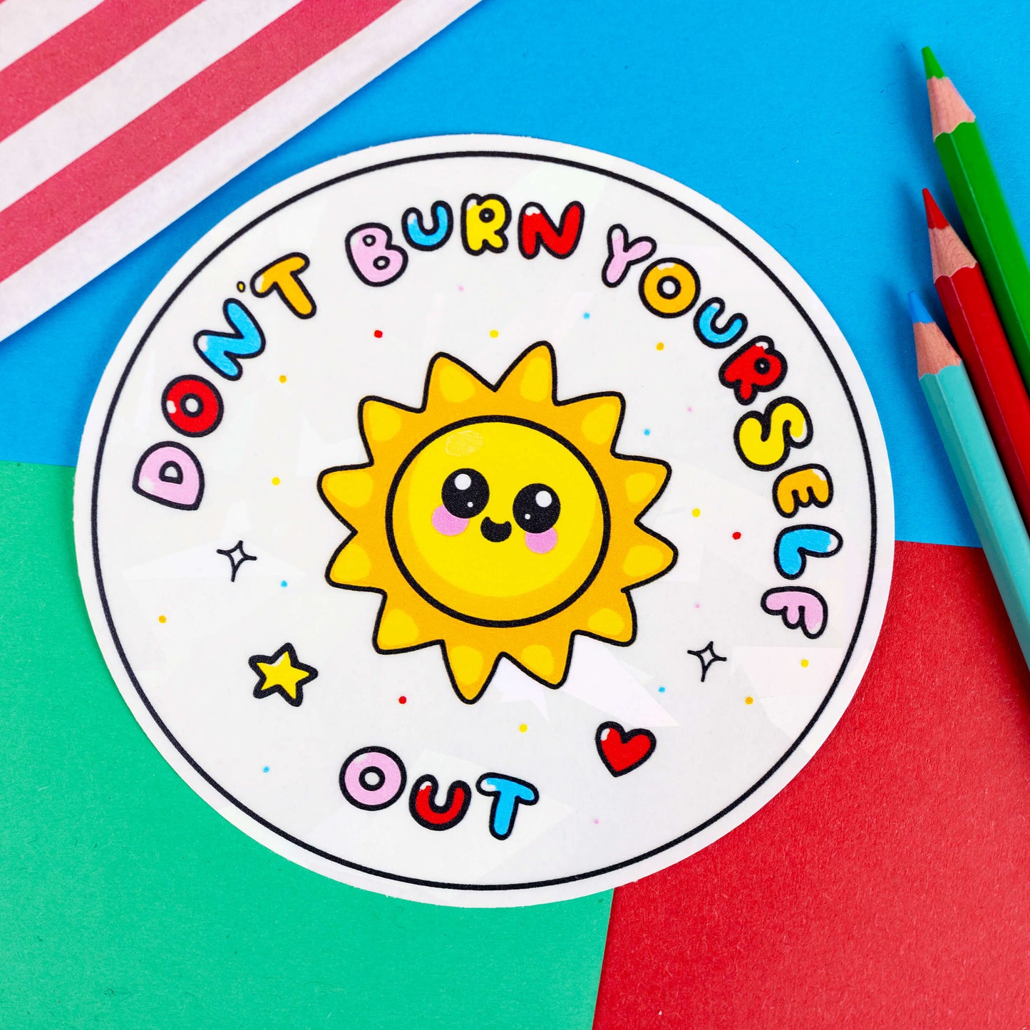 The Don't Burn Yourself Out Sun Catcher Rainbow Window Sticker on a red, blue and green background with colouring pencils and red stripe candy bag. The circle holographic sticker features a smiling sunshine with pink blush in the middle with rainbow bubble writing reading 'don't burn yourself out' with a yellow star, red heart, black sparkle outlines and rainbow dots all over. The design is inspired by self care.