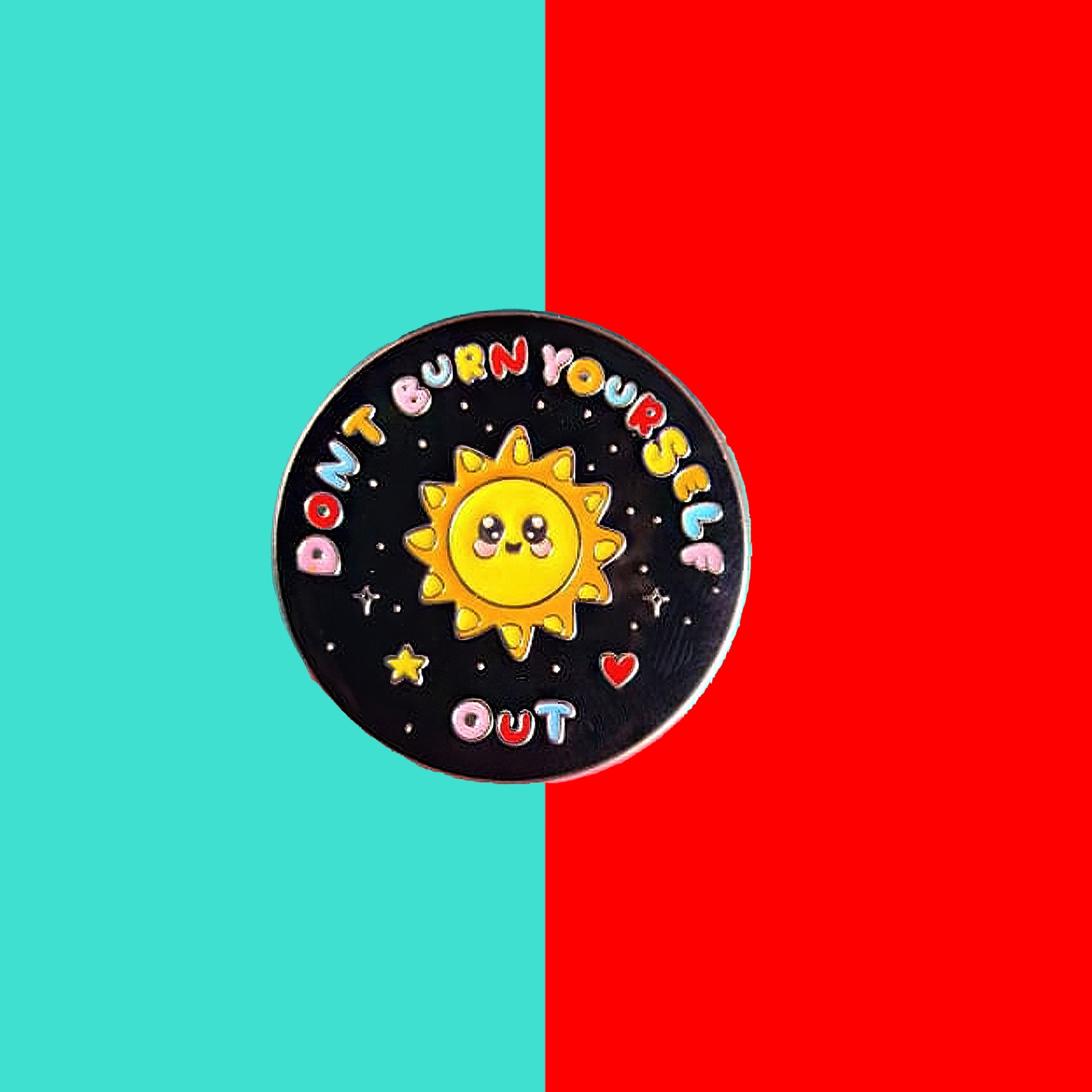 The Don't Burn Yourself Out Sunshine Enamel Pin on a blue and red background. The circle shaped pin badge is a black base with a yellow smiling sunshine in the centre with rainbow text surrounding it reading 'don't burn yourself out' along with a red heart, yellow star and silver sparkles. The design was created as a gentle reminder to take care of yourself and self care. 