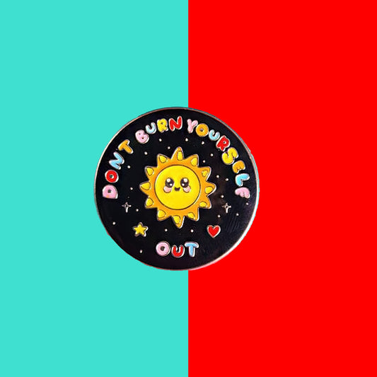 The Don't Burn Yourself Out Sunshine Enamel Pin on a blue and red background. The circle shaped pin badge is a black base with a yellow smiling sunshine in the centre with rainbow text surrounding it reading 'don't burn yourself out' along with a red heart, yellow star and silver sparkles. The design was created as a gentle reminder to take care of yourself and self care. 