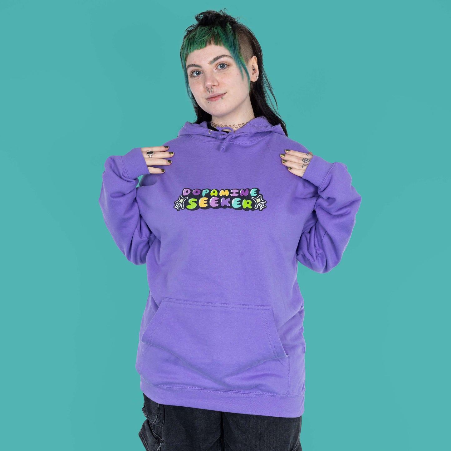 The Dopamine Seeker Hoodie in digital lavender being modelled by Faeryn who has a green and black mullet, they have styled the cosy hoodie with wide leg charcoal jeans. They are facing forward smiling with both hands resting on their shoulders. The hoodie design has 'dopamine seeker' written in the middle in rainbow bubble font and black sparkle outlines. The hoodie has purple drawstrings and a large centre pocket. Design is raising awareness for ADHD and neurodivergence.