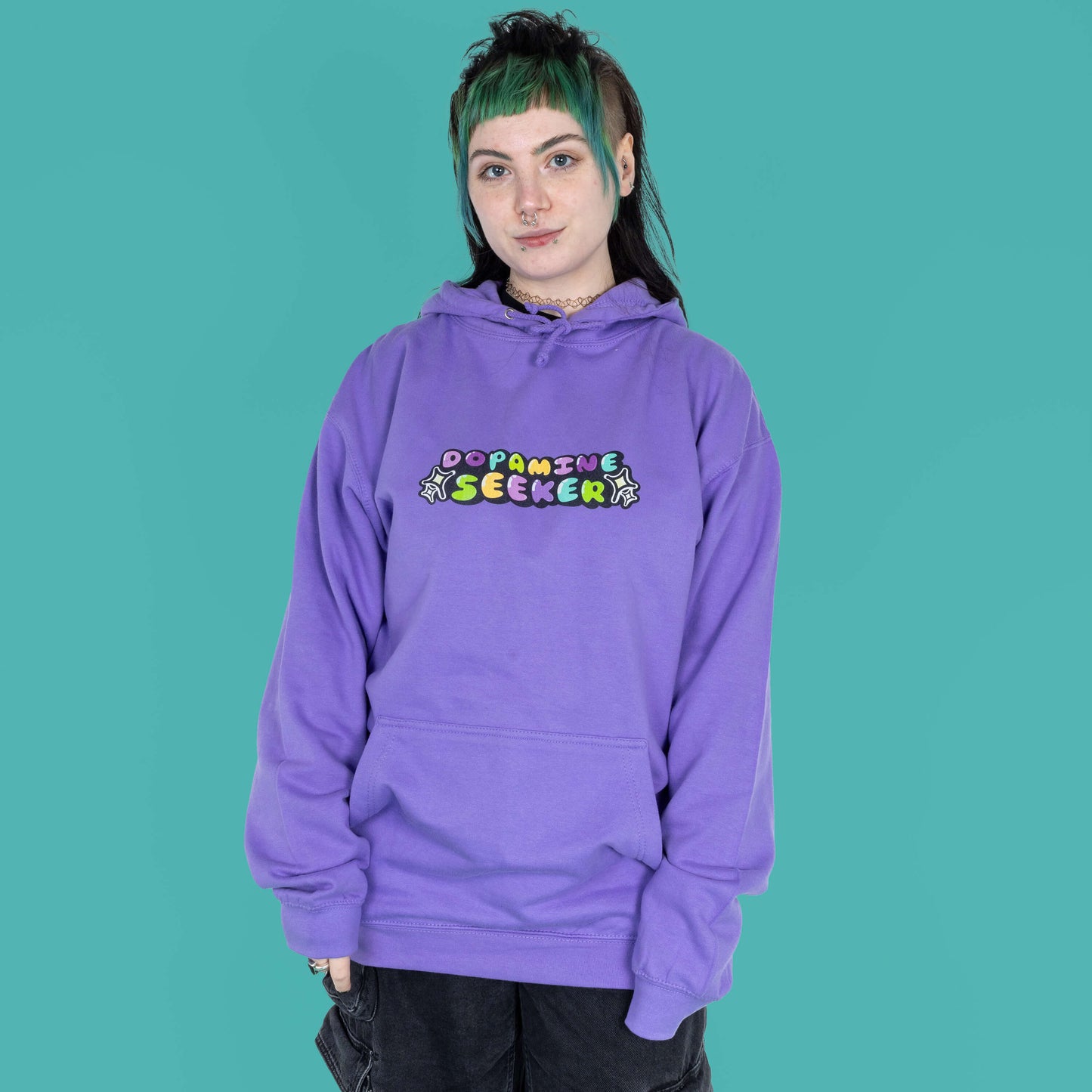 The Dopamine Seeker Hoodie in digital lavender being modelled by Faeryn who has a green and black mullet, they have styled the cosy hoodie with wide leg charcoal jeans. They are facing forward smiling with their hands by their side. The hoodie design has 'dopamine seeker' written in the middle in rainbow bubble font and black sparkle outlines. The hoodie has purple drawstrings and a large centre pocket. Design is raising awareness for ADHD and neurodivergence.