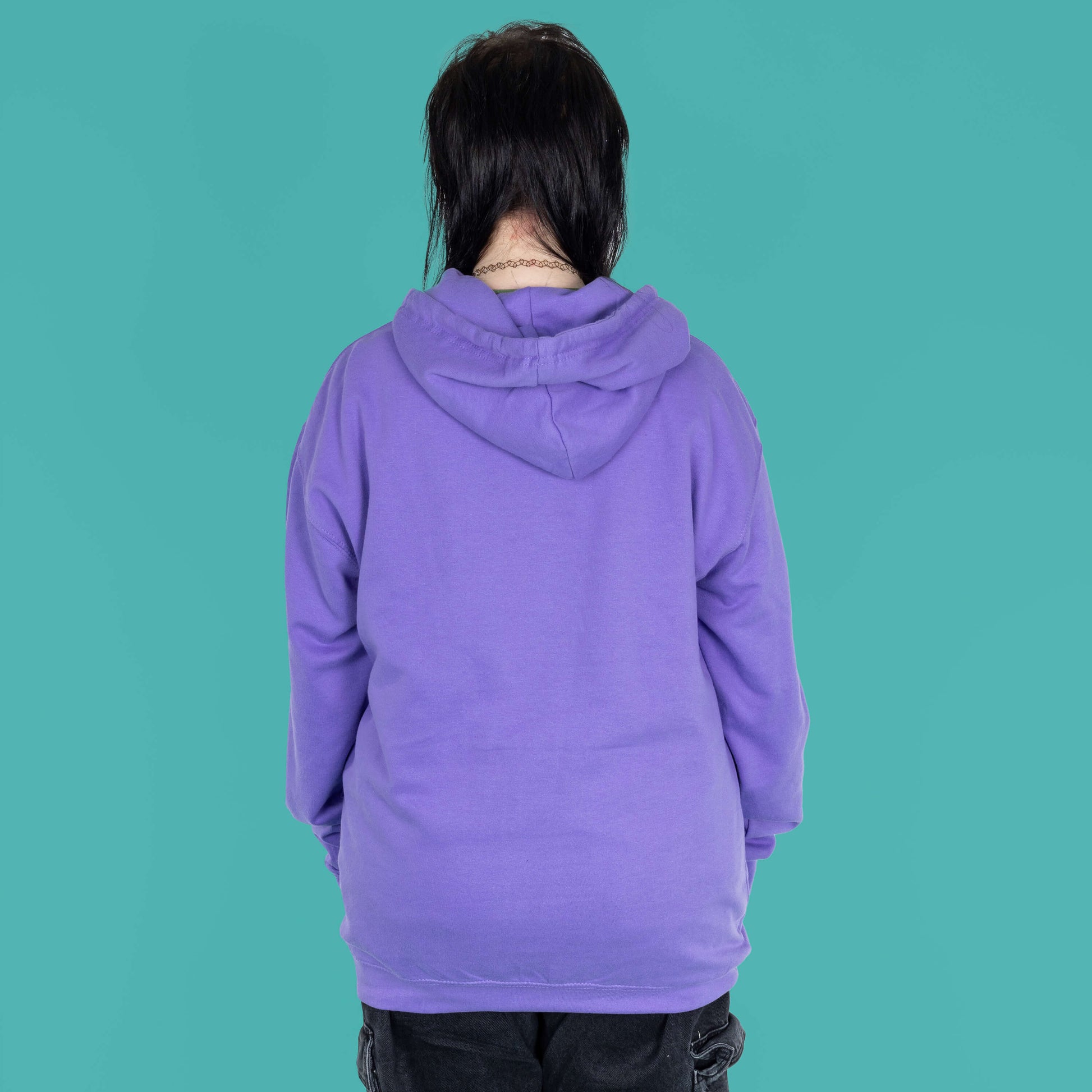 The Dopamine Seeker Hoodie in digital lavender being modelled by Faeryn who has a green and black mullet, they have styled the cosy hoodie with wide leg charcoal jeans. They are facing away from camera to show the plain pastel purple back. The hoodie design has 'dopamine seeker' written in the middle in rainbow bubble font and black sparkle outlines. The hoodie has purple drawstrings and a large centre pocket. Design is raising awareness for ADHD and neurodivergence.