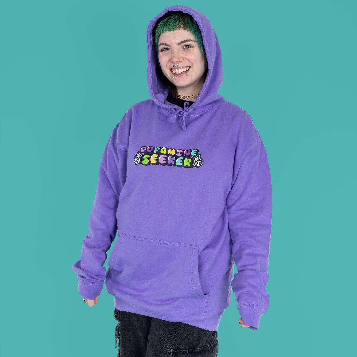 The Dopamine Seeker Hoodie in digital lavender being modelled by Faeryn who has a green and black mullet, they have styled the cosy hoodie with wide leg charcoal jeans. They are facing forward smiling with the hood up and their hands by their side. The hoodie design has 'dopamine seeker' written in the middle in rainbow bubble font and black sparkle outlines. The hoodie has purple drawstrings and a large centre pocket. Design is raising awareness for ADHD and neurodivergence.