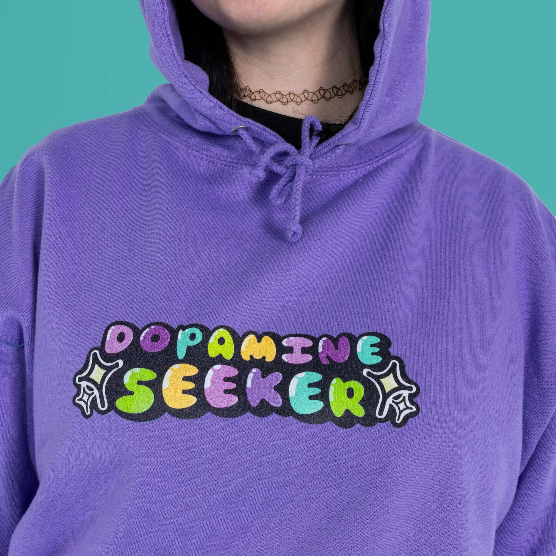 Close up of The Dopamine Seeker Hoodie in digital lavender being modelled by Faeryn. The hoodie design has 'dopamine seeker' written in the middle in rainbow bubble font and black sparkle outlines. The hoodie has purple drawstrings and a large centre pocket. Design is raising awareness for ADHD and neurodivergence.