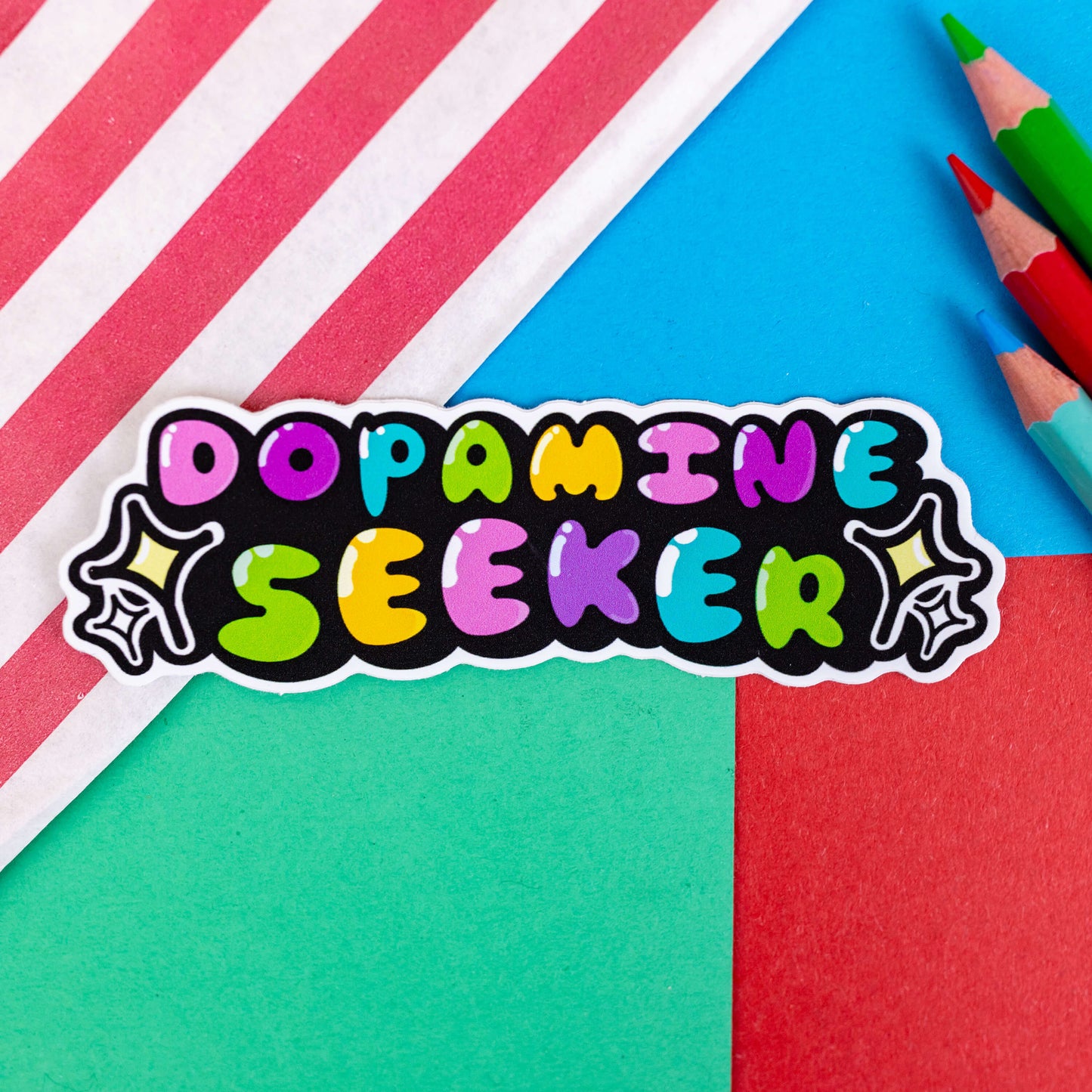 The Dopamine Seeker Sticker on a red, blue and green background with colouring pencils and a red stripe candy bag. The rainbow bubble font sticker reads 'dopamine seeker' with yellow, black and white sparkles either side. The design is raising awareness for ADHD and neurodivergence.