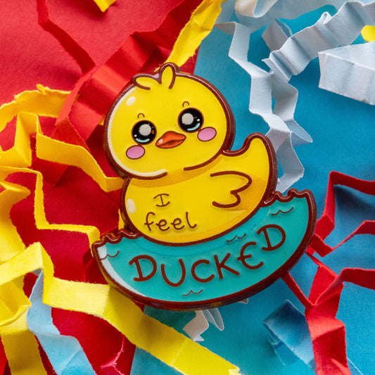 Close up of the Innabox I Feel Ducked enamel pin featuring a cute yellow duck with rosy cheeks sitting in blue water. The hard enamel pin has glossy detailing and playful text reading I feel ducked. Bright red, yellow and blue paper confetti background.