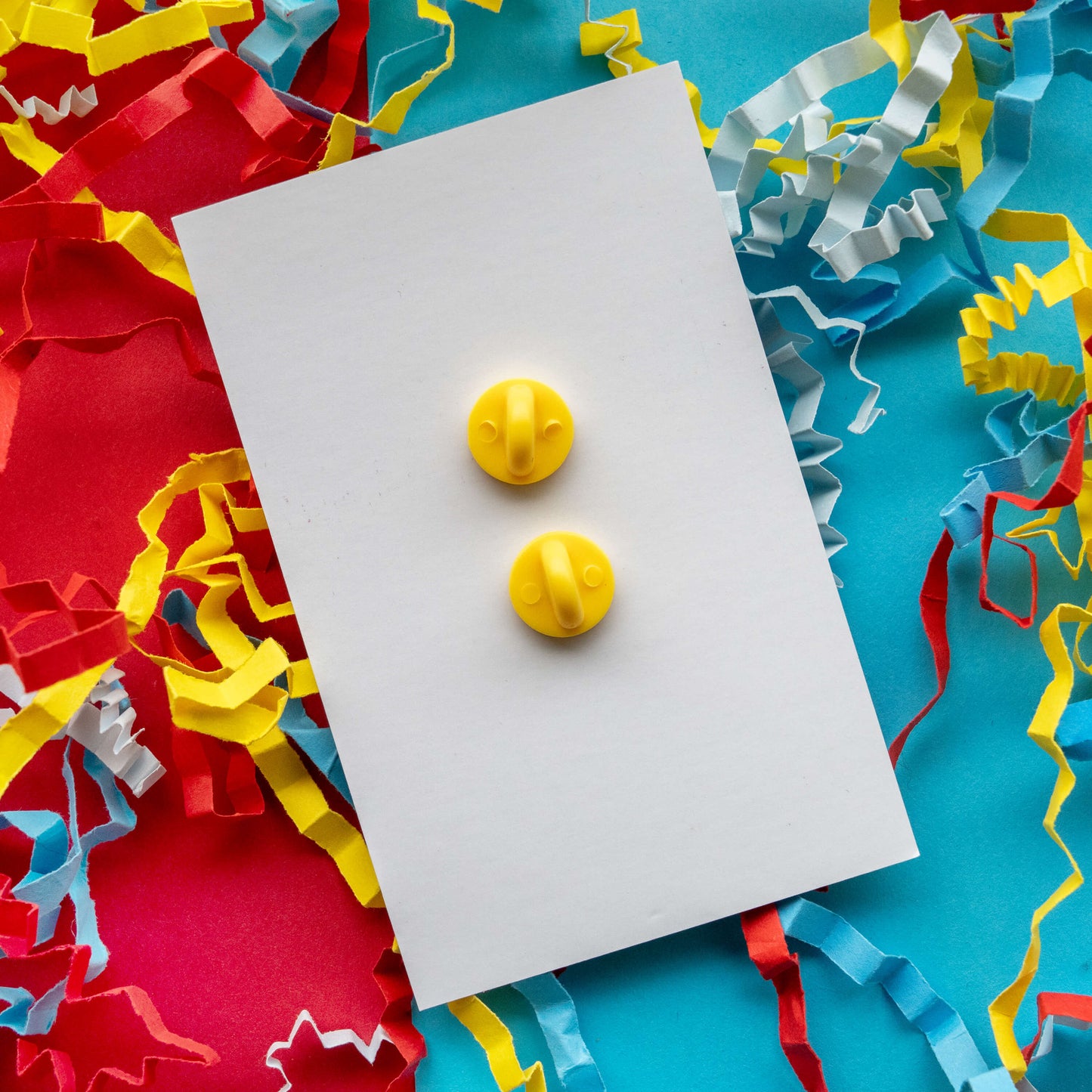 Back view of the Innabox I Feel Ducked enamel pin showing two yellow rubber clutch backs on a white display card. The disability inspired hard enamel pin is photographed on a colourful red and blue background with curled paper streamers.