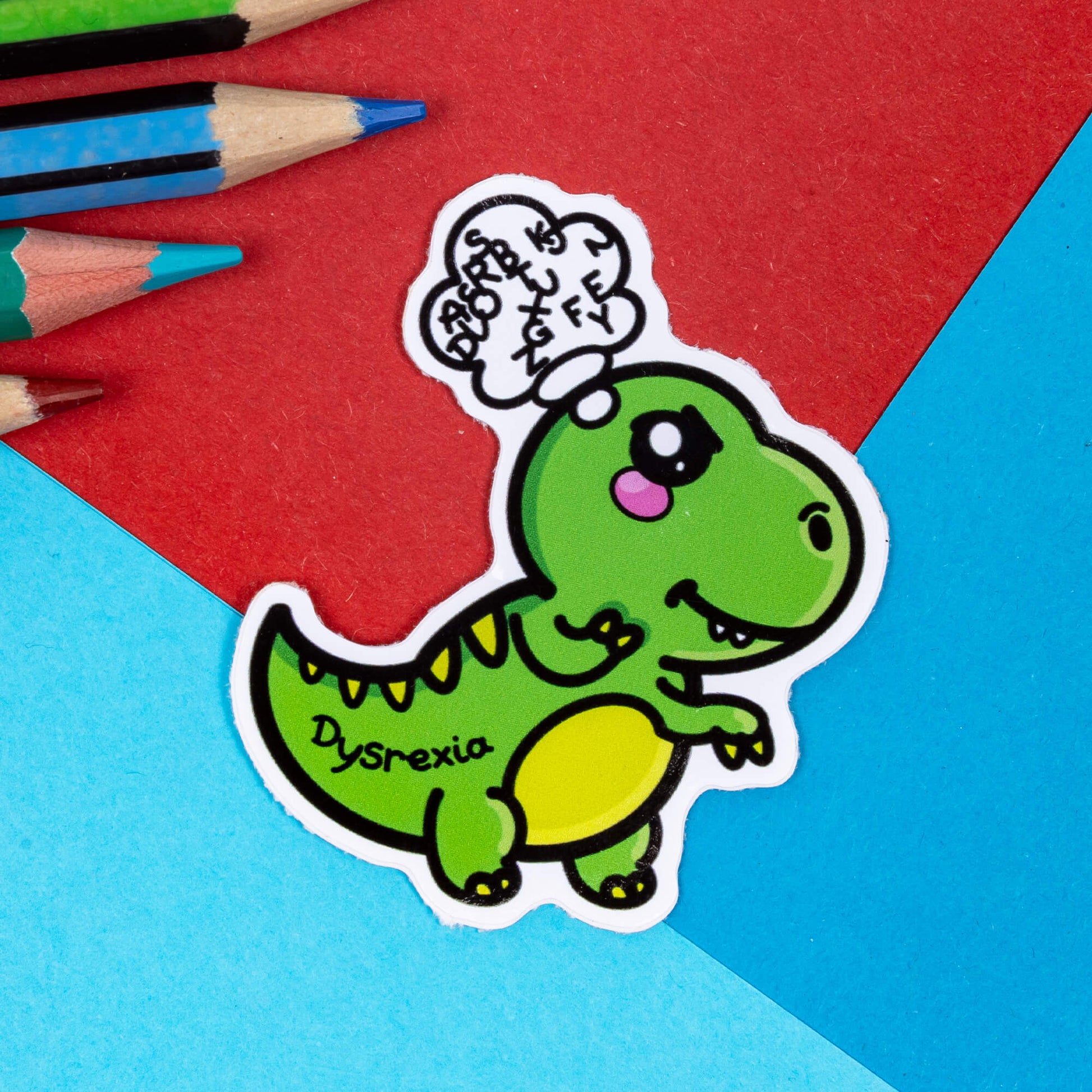 A sticker of a little green dinosaur with Dysrexia written on their tail and muddled letters in a speech bubble above their head on a red and blue background