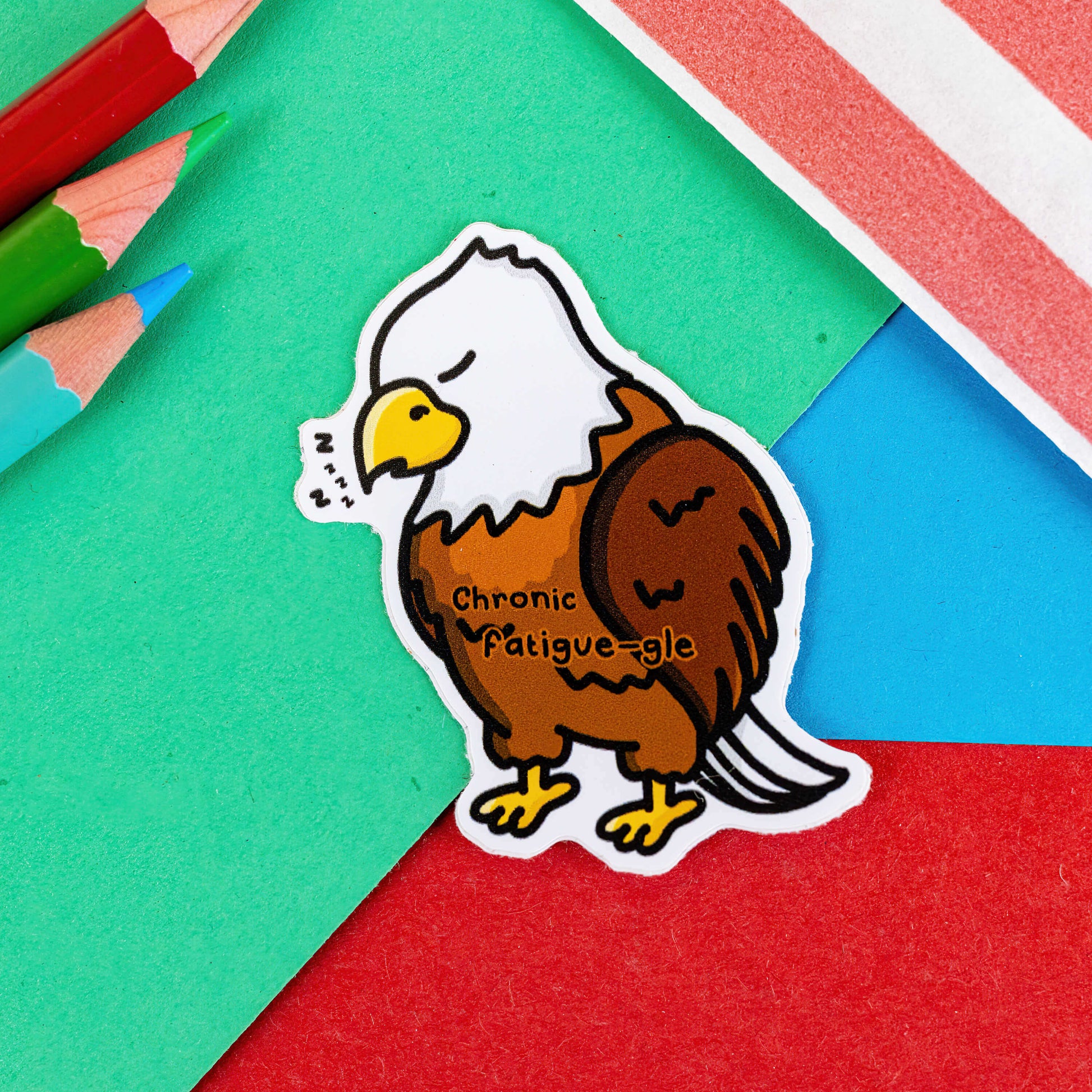The Chronic Fatigue-le Sticker - Chronic Fatigue on a red, blue and green background with colouring pencils and a red stripe candy bag. The sticker is a sleeping tired brown and white eagle with a yellow beak and feet, across the middle in black text reads 'chronic fatigue-gle'. The design was created to raise awareness for chronic fatigue / ME / CFS.