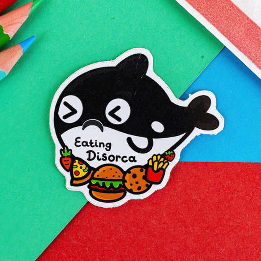 The Eating Disorca Orca Whale Sticker - Eating Disorder on a red, blue and green background with colouring pencils and a red stripe candy bag. The black orca whale shaped vinyl sticker has a stressed expression whilst surrounded by burgers, pizzas, cookies, fries and fruit with 'eating disorca' written across its middle. The design is raising awareness for eating disorders.