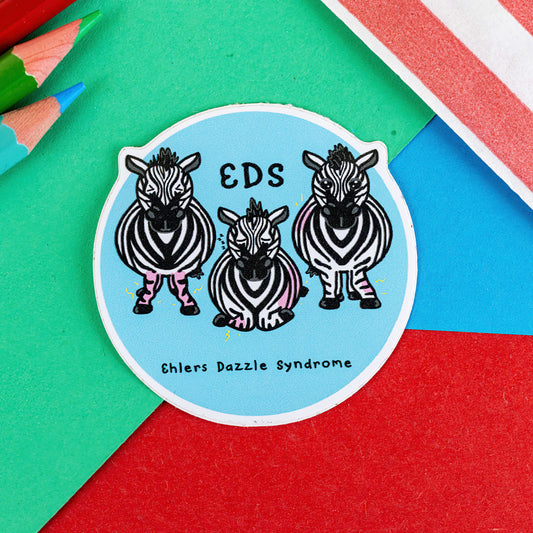 A sticker of a blue circle with three swollen and in pain zebras with the letters EDS above and Ehlers Dazzle Syndrome written on the bottom it is on a green, red and blue background with pencils in in the corner of the shot. The hand drawn sticker design is raising awareness for Ehlers-Danlos Syndrome EDS.