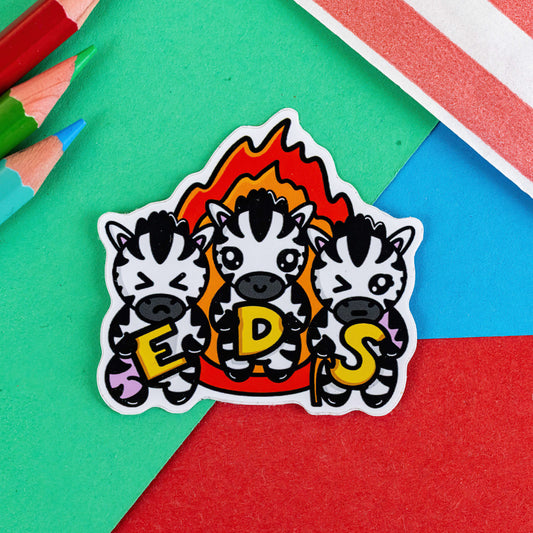 A sticker with three zebras on with a flame behind them and they are holding letters spelling EDS it is on a green, red and blue background with pencils in in the corner of the shot. Hand drawn design is raising awareness for Ehlers-Danlos Syndrome.