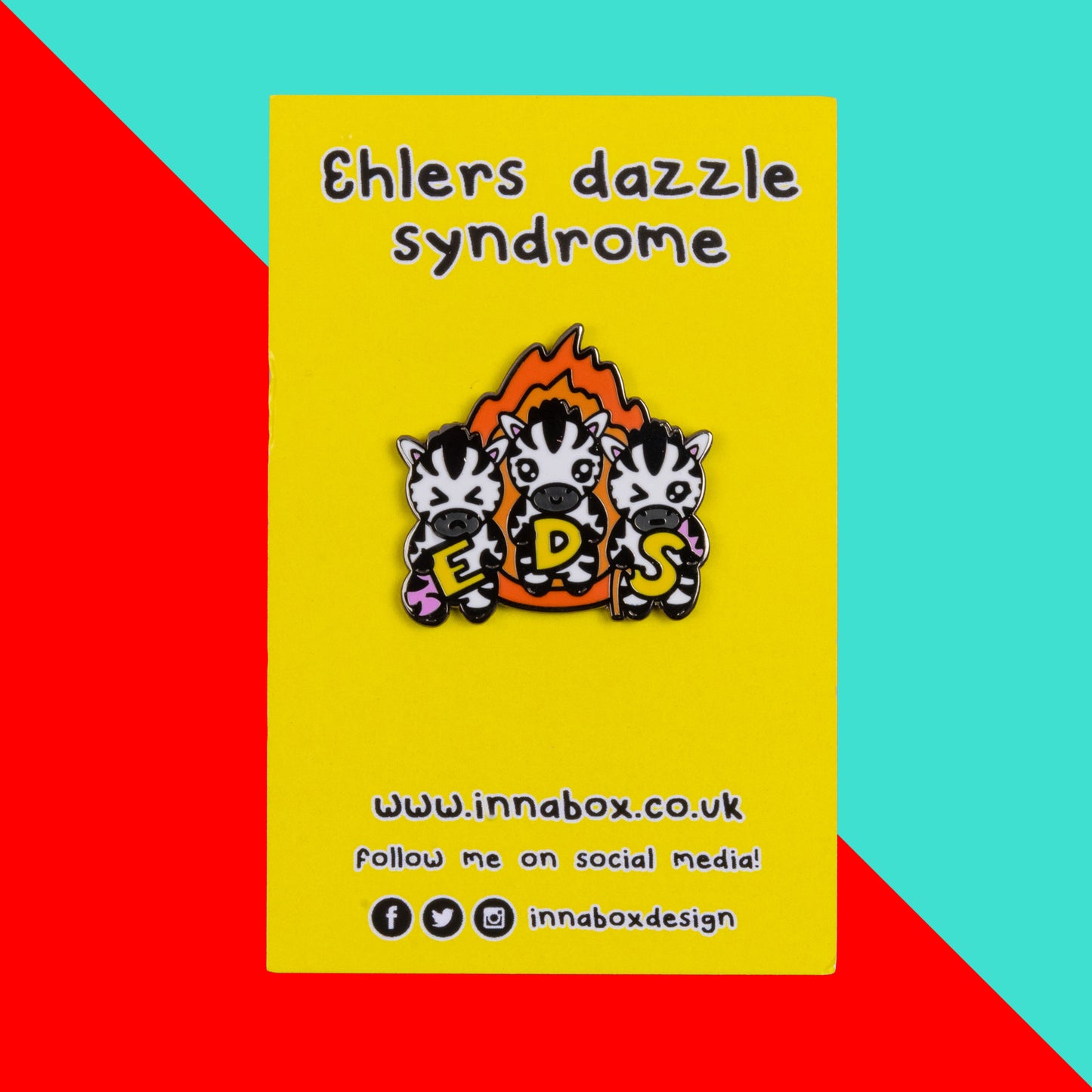 The Ehlers Dazzle Syndrome EDS Enamel Pin 2.0 - Ehlers-Danlos Syndrome on yellow backing card with black text above reading 'ehlers dazzle syndrome' and below reading the innabox website and social media handles on a red and blue background. A pin of three zebras in front of an orange flame holding up yellow letters spelling out EDS, the first zebra has a swollen pink leg, the next is smiling and the third has a swollen pink arm and a walking stick. Raising awareness for Ehlers-Danlos Syndrome.