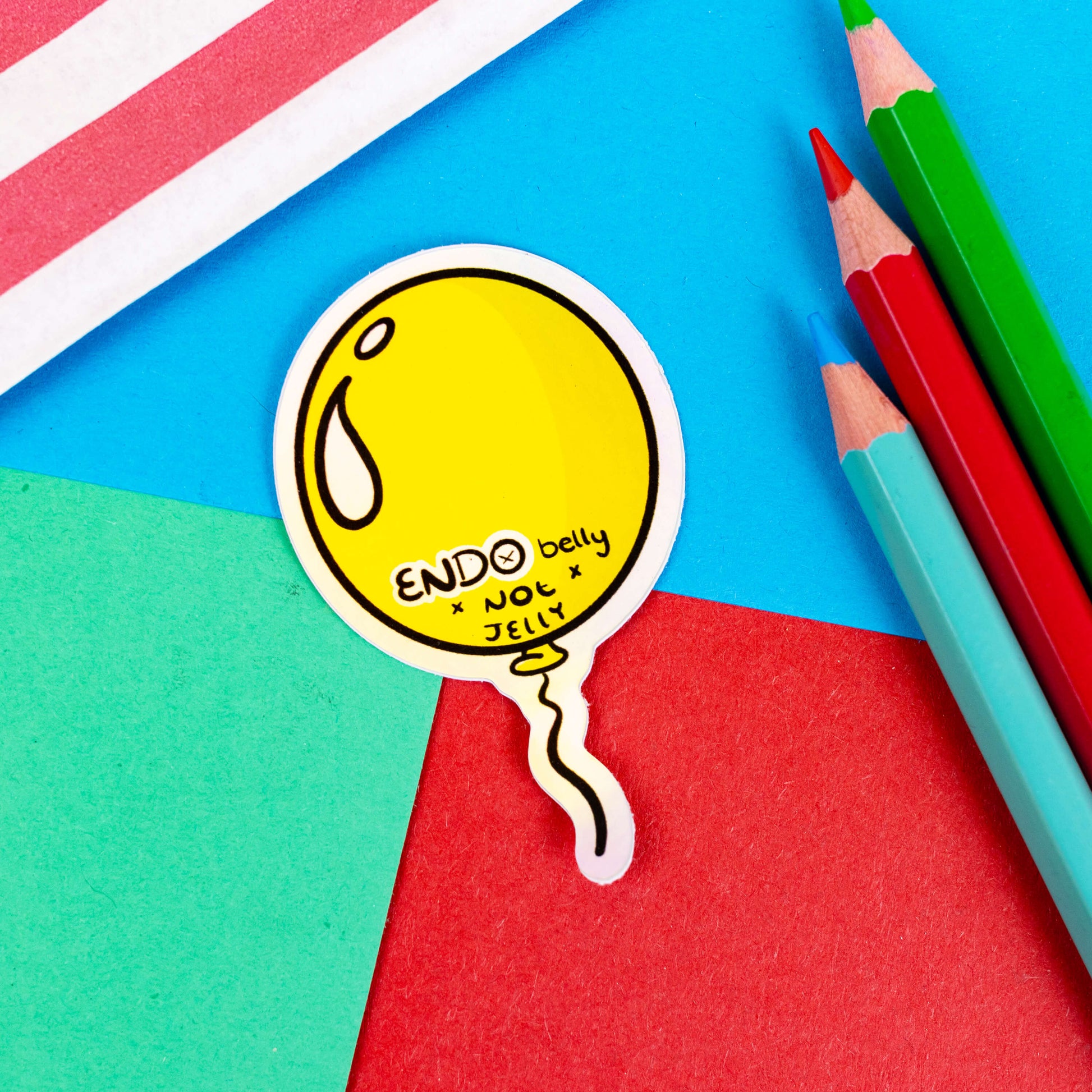 The Endo Belly Sticker on a red, blue and green background with colouring pencils and red stripe candy bag. The yellow balloon shape sticker has bottom black text reading 'ENDO belly not jelly'. The hand drawn design is raising awareness for endometriosis.