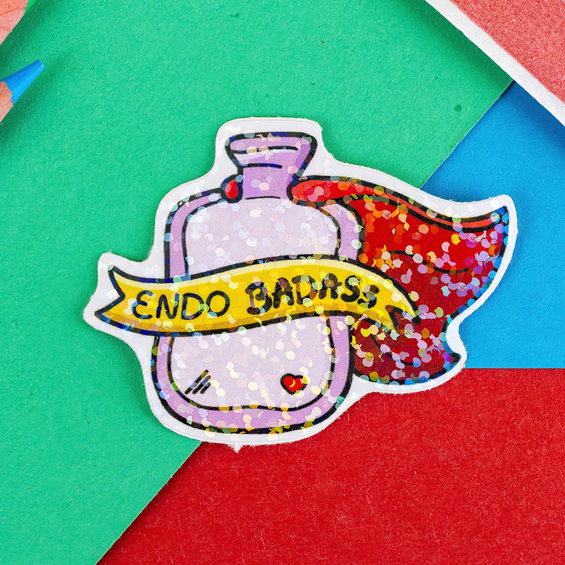 The Endo Badass Sticker - Endometriosis on a red, blue and green background with colouring pencils and red stripe candy bag. The holographic shiny sticker is of a pink hot waterbottle with a red small heart, a red cape and yellow banner going across with black text reading 'Endo Badass'. Hand drawn design is raising awareness for endometriosis and pelvic pain.